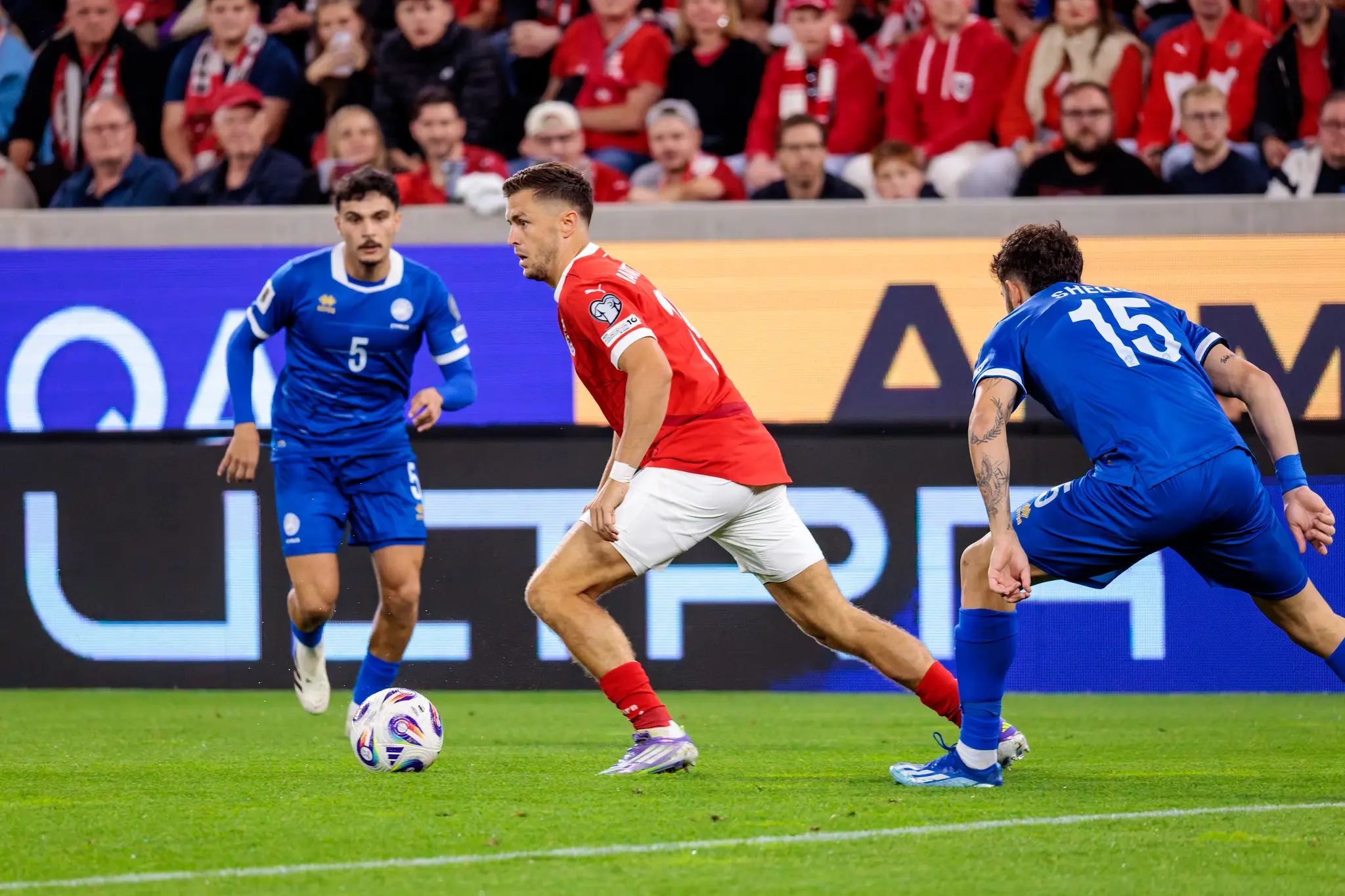 Fußballspieler Christoph Baumgartner im roten Trikot dribbelt den Ball im Zweikampf mit zwei zyprischen Spielern in blauen Trikots während eines Länderspiels