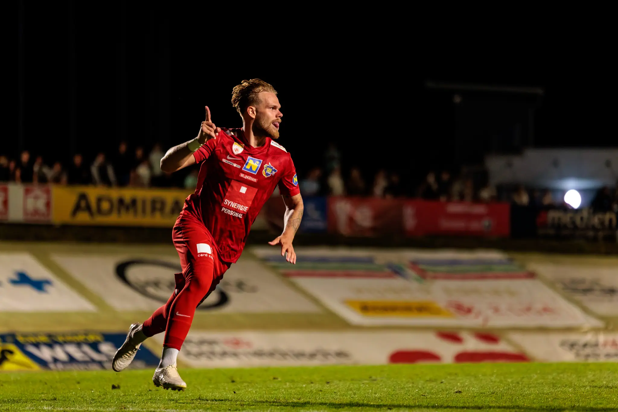 Ein Fußballspieler des SKN St. Pölten in roter Spielkleidung feiert auf dem nächtlichen Spielfeld ausgelassen einen Erfolg mit erhobenem Finger