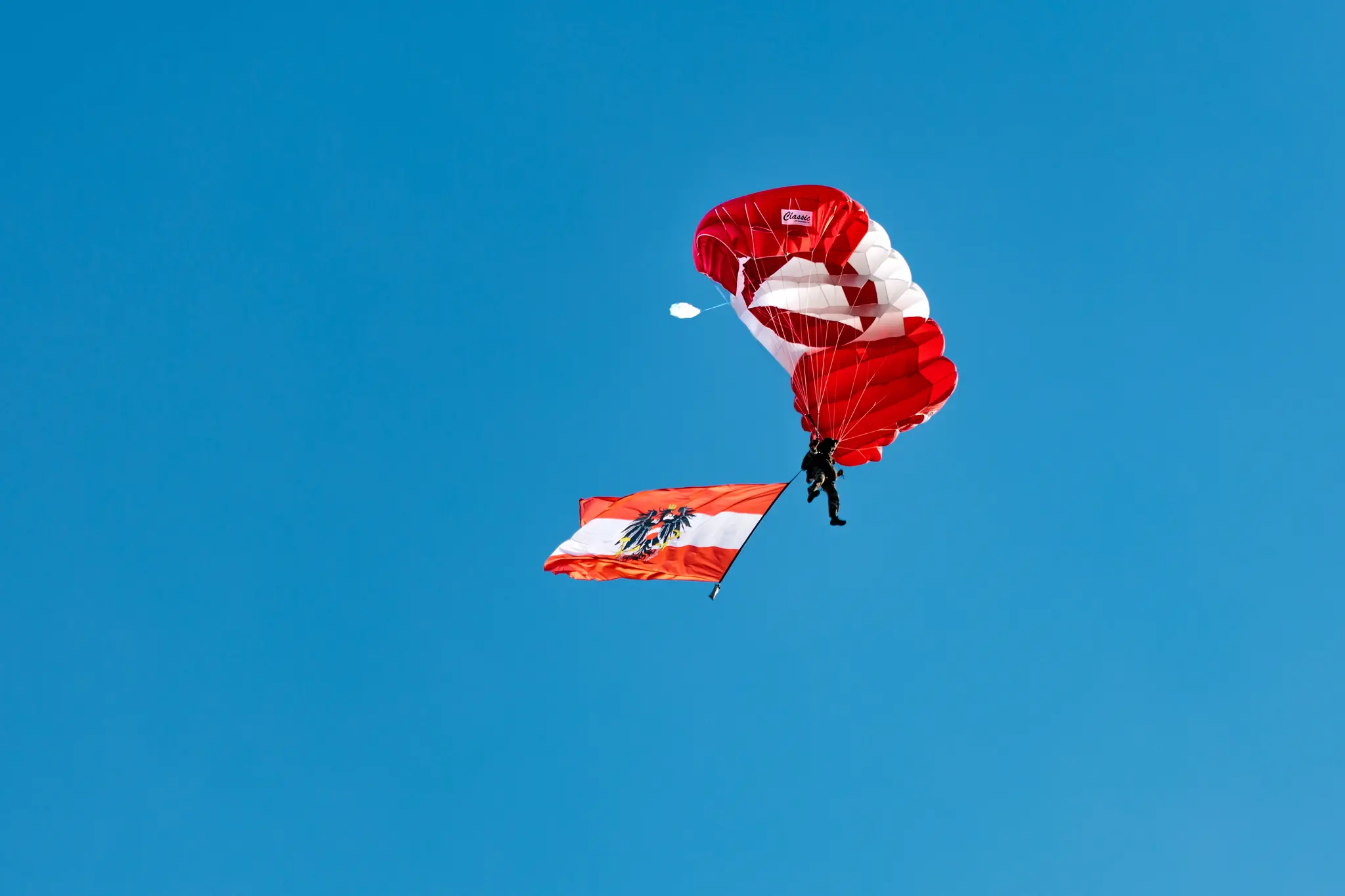 Ein Bundesheer-Springer schwebt noch in der Luft unter rot-weiß-rotem Fallschirm, darunter weht eine österreichische Flagge.