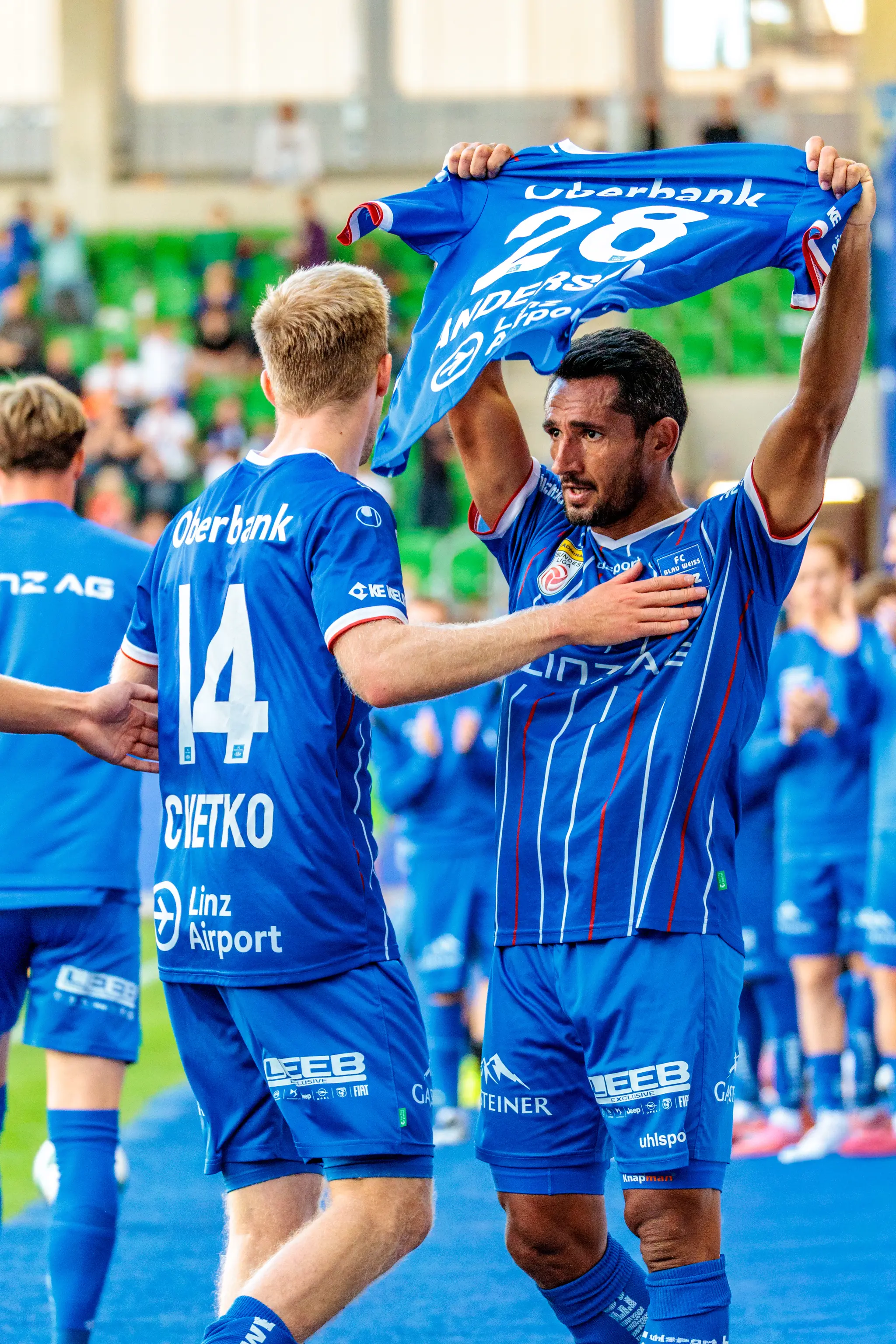 Der Fußballspieler Ronivaldo vom FC Blau-Weiß Linz hält nach seinem Tor emotional das Trikot des verletzten Anders Capek hoch, während sein Teamkollege Christopher Cvetko mit ihm feiert