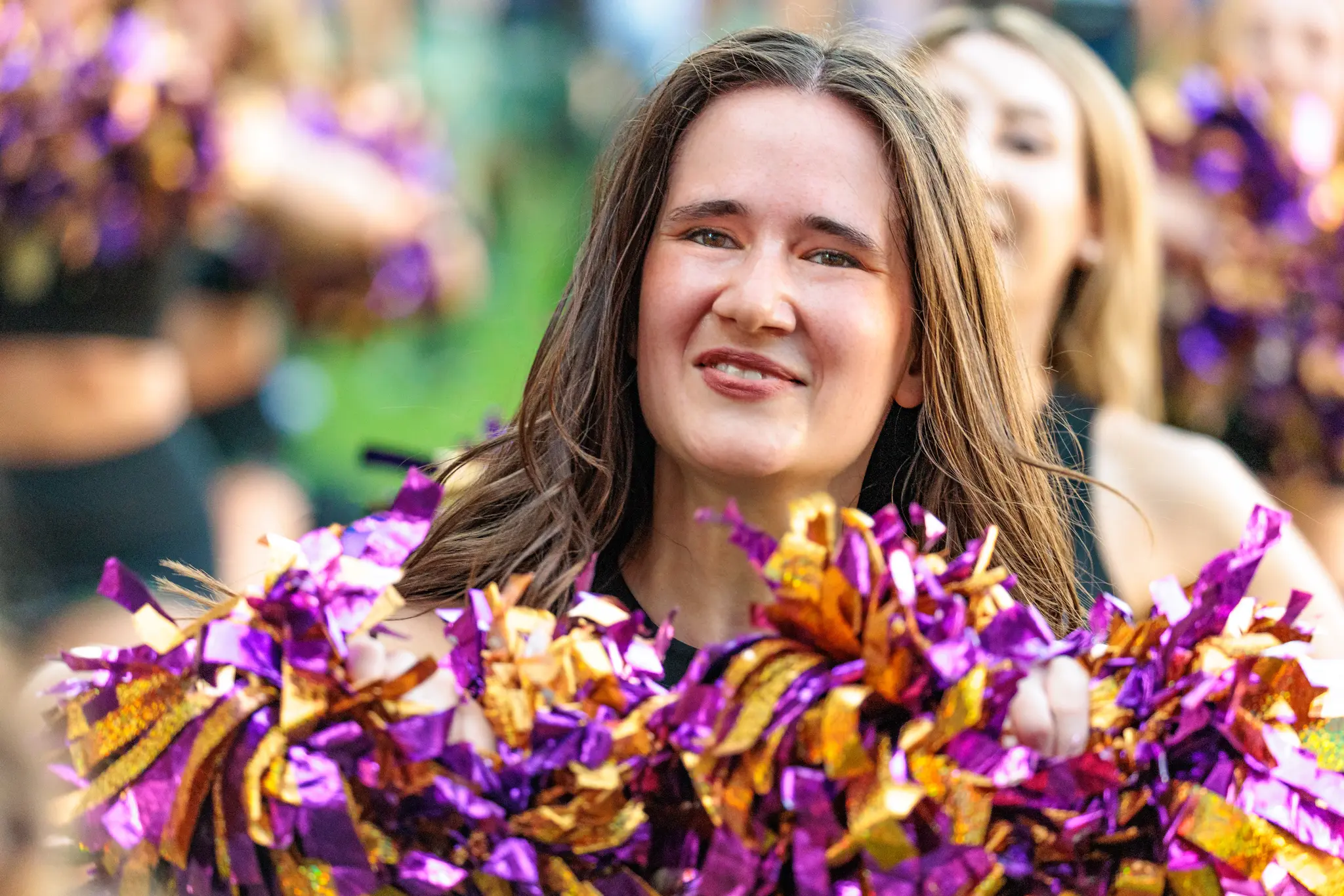 Porträt einer Cheerleaderin des Spirit Squad mit violett-goldenen Pompons vor unscharfer Stadionkulisse.