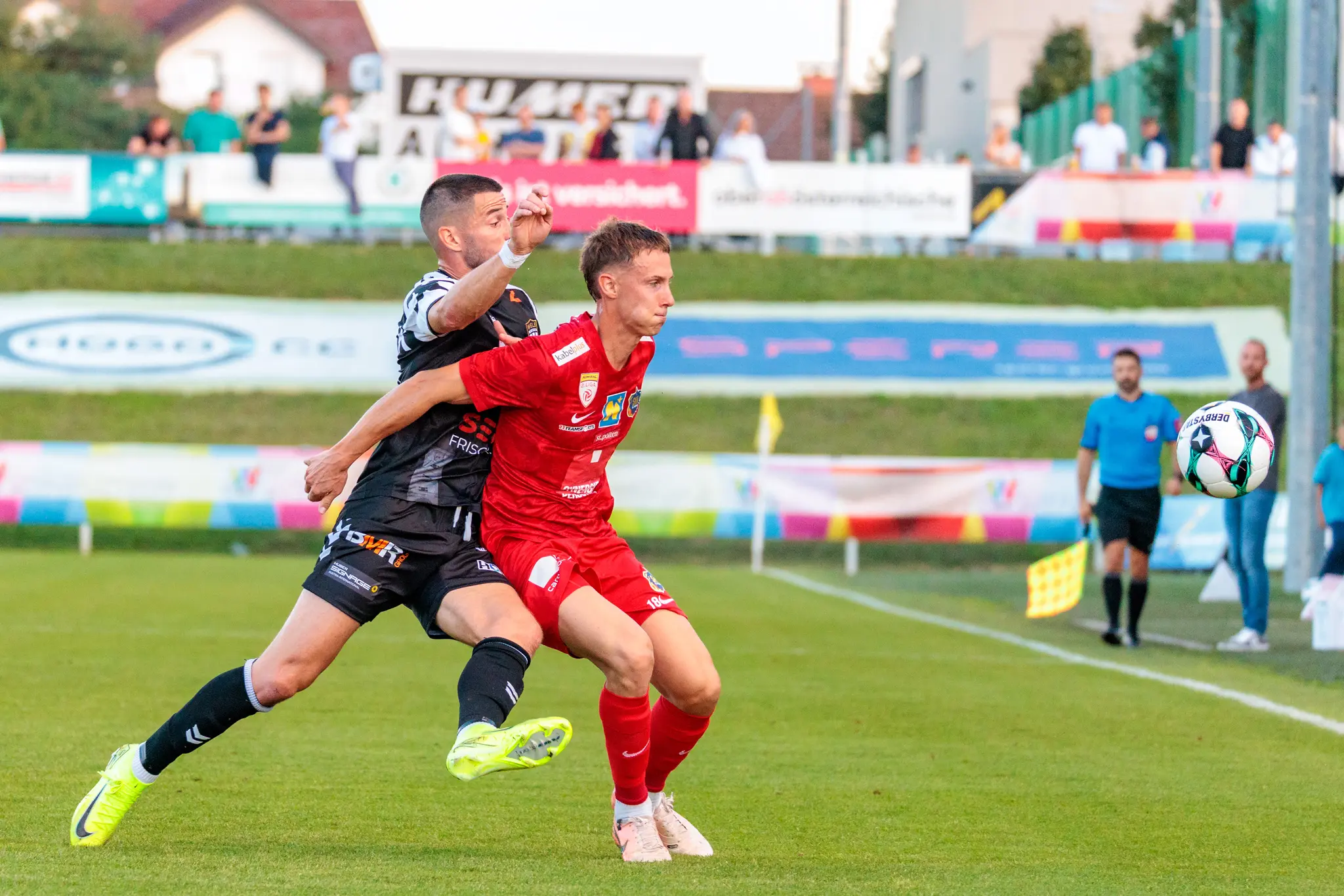 Ein Spieler in rotem Trikot und ein Spieler in schwarz-weißem Trikot ringen auf dem Spielfeld energisch um den Fußball
