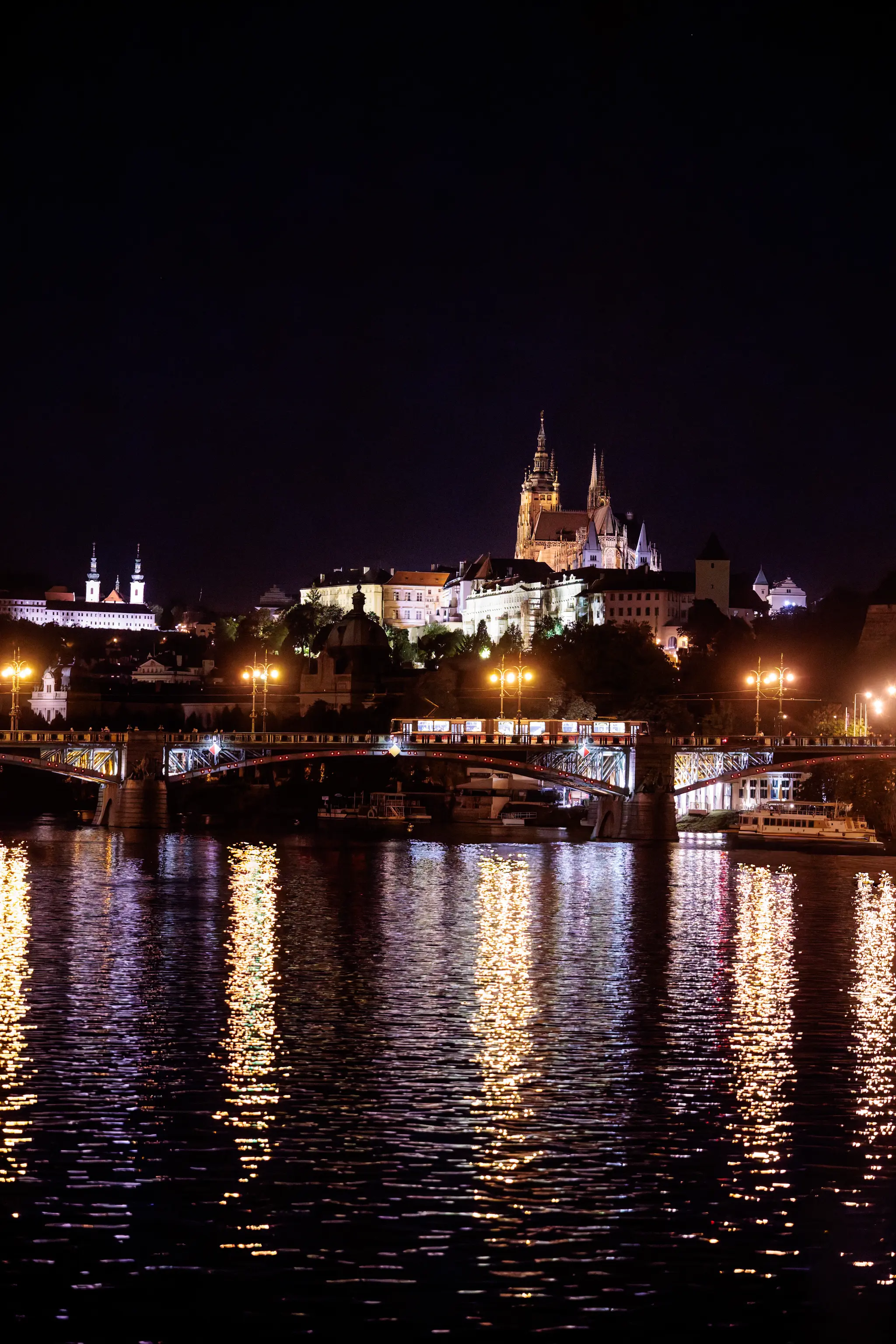 Die beleuchtete Prager Burg mit dem Veitsdom über der Moldau, während eine Straßenbahn über die Brücke fährt und sich die Lichter im Fluss spiegeln.