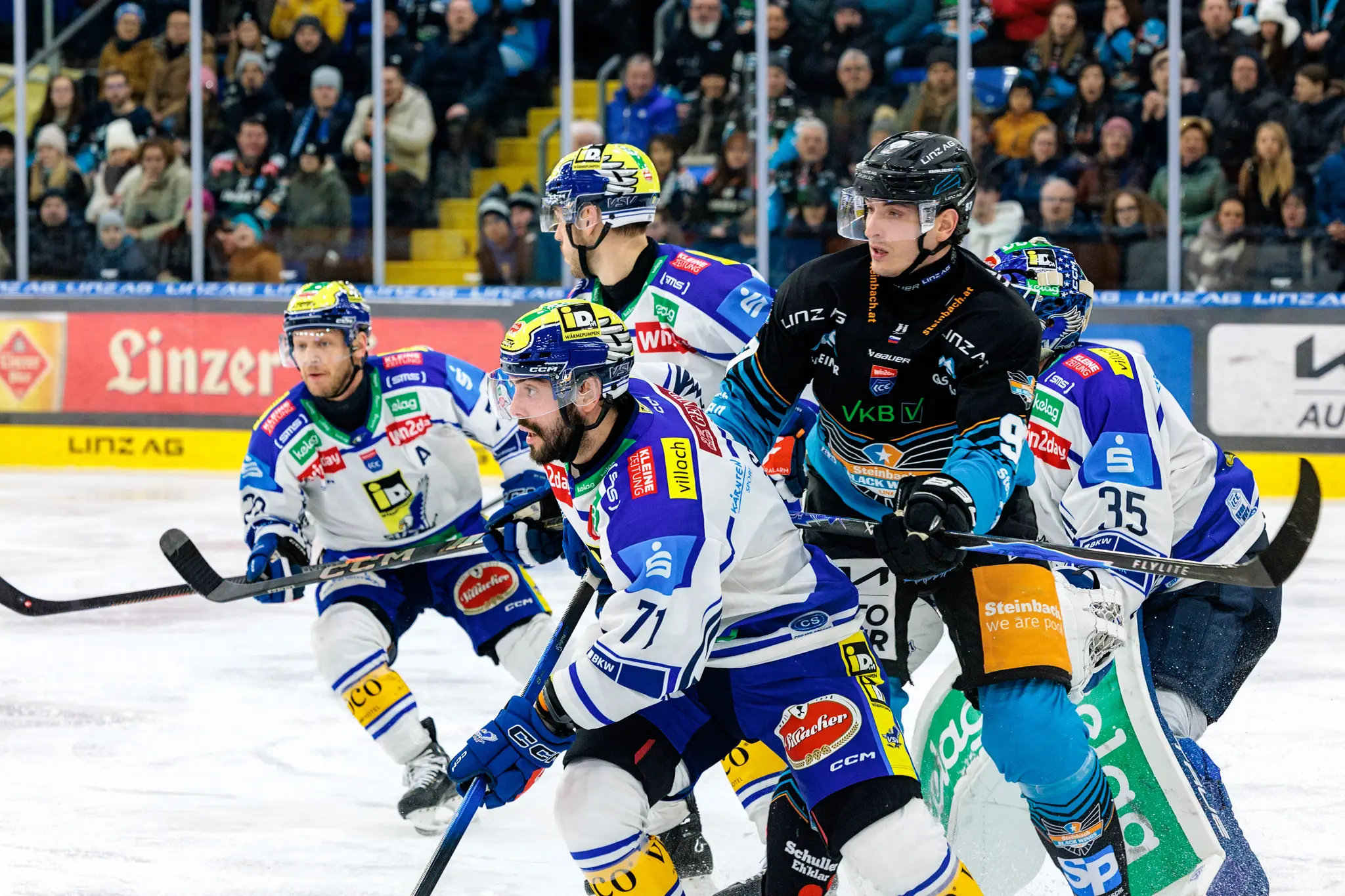 Eishockeyspieler der Black Wings Linz in schwarzen Trikots und des VSV in blauen Trikots kämpfen intensiv um den Puck auf dem Eis, während der Torhüter des VSV bereitsteht