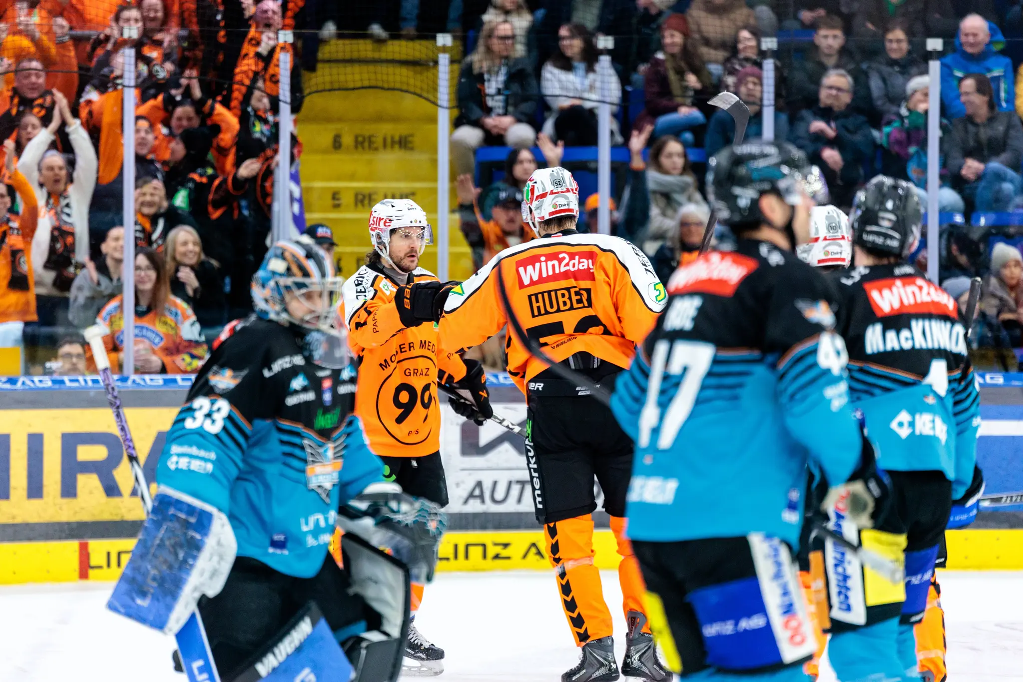 Eishockeyspieler der Grazer 99ers in orangefarbenen Trikots feiern das 1:0 auf dem Eis, während im Hintergrund die grazer Fans jubeln und die Fans der BlackWings eher betrübt aussehen