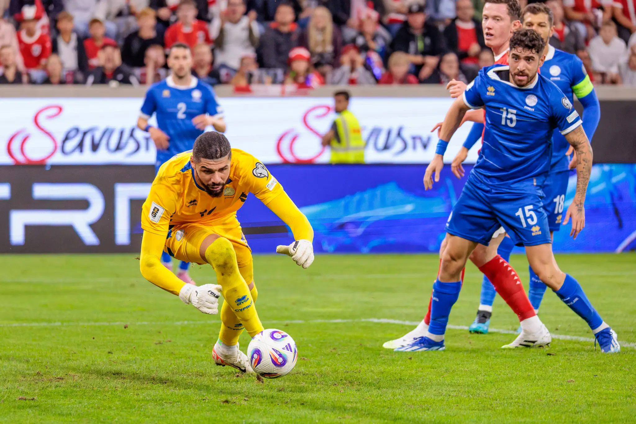 Der zypriotische Torhüter Fabiano im gelben Trikot kontrolliert den Ball am Boden, während seine Teamkollegen Christos Shelis (Nummer 15) und Kostas Pileas (Nummer 2) im Hintergrund positioniert sind