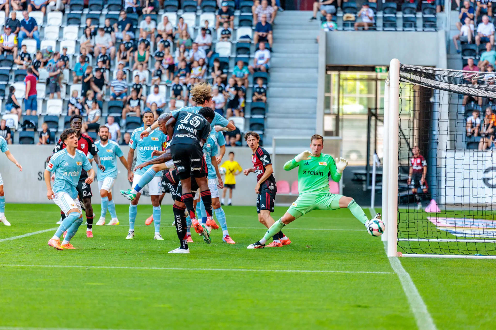 Ein grüner Torhüter hält in der Raiffeisen Arena im Sprung einen Ball vom Tor fern, umgeben von Spielern in hellblauen und dunklen Trikots 