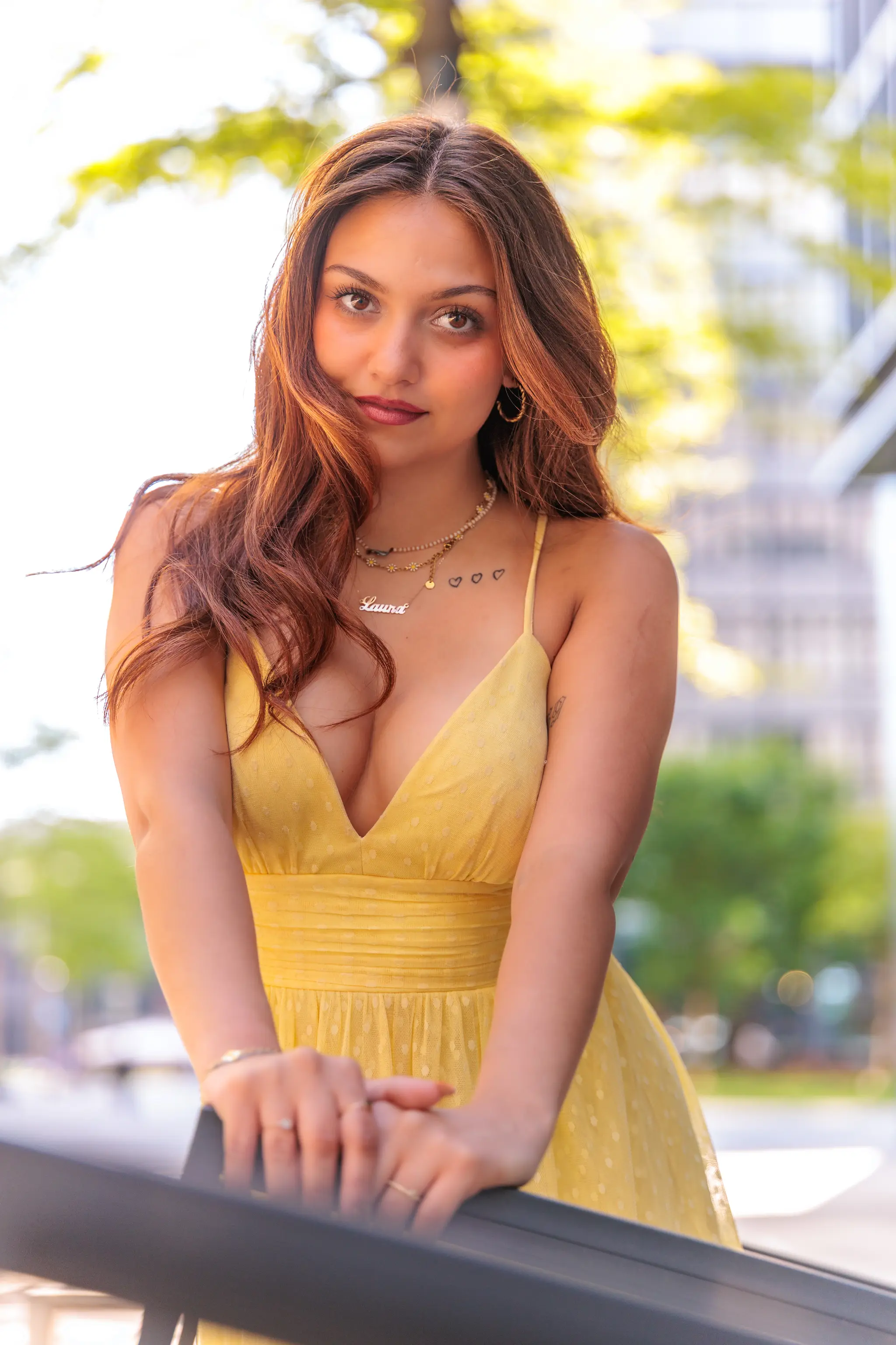 Porträt einer jungen Frau mit langen braunen Haaren in einem gelben Sommerkleid, die lächelnd in die Kamera blickt, vor einem verschwommenen urbanen Hintergrund bei Tageslicht. 