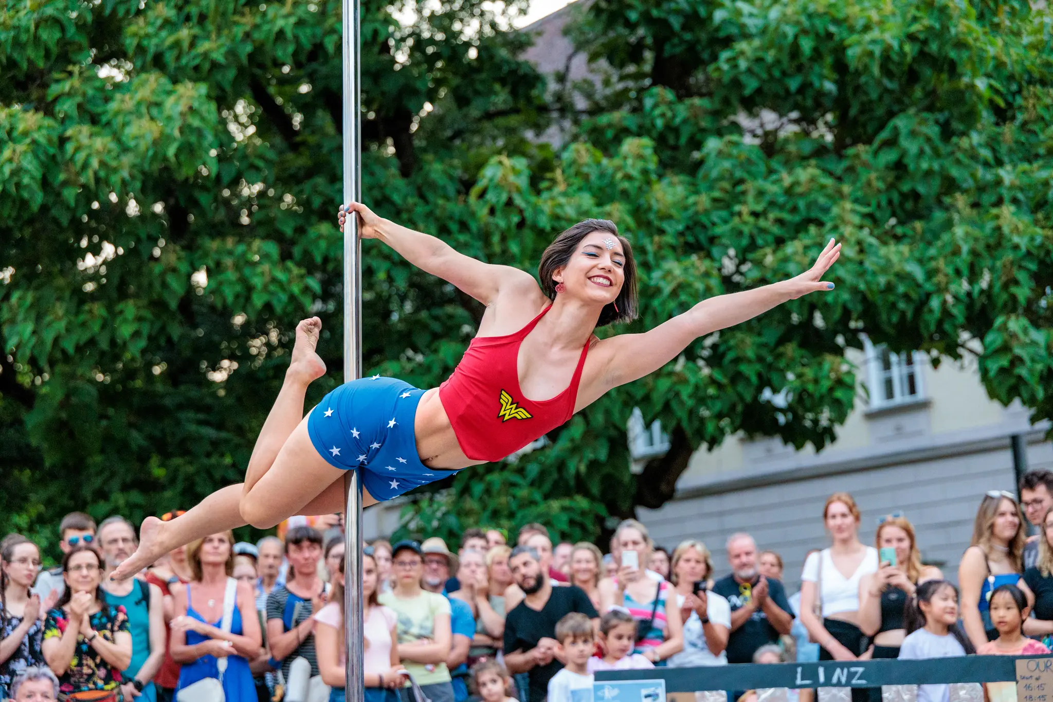 Lachende Frau in Wonder Woman-Outfit beim Pole Dance Show während des Pflasterspektakels in LInz, in akrobatischer Pose vor einem Publikum und Bäumen 