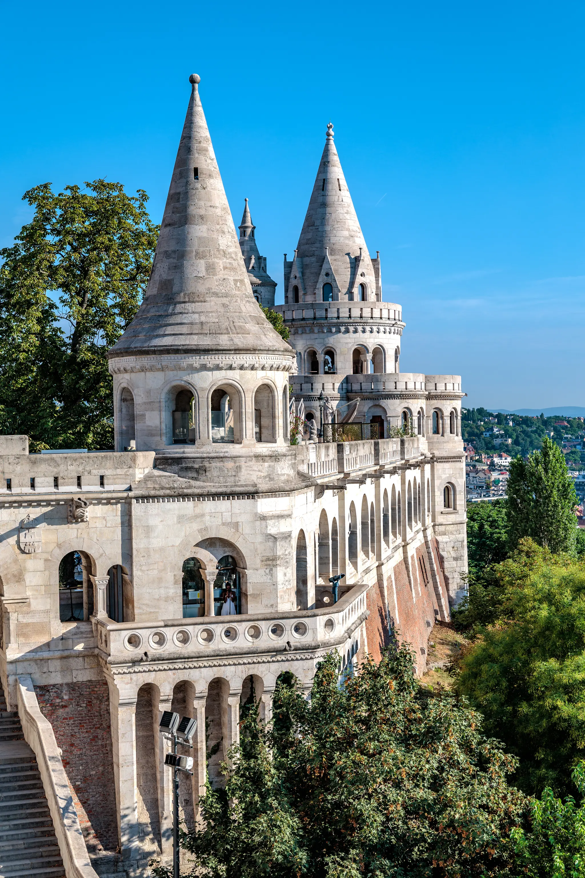 Majestätische Fischerbastei in Budapest mit weißen, spitz zulaufenden Steintürmen, zahlreichen Arkaden und Balustraden, umgeben von grünen Bäumen unter klarem blauem Himmel. 