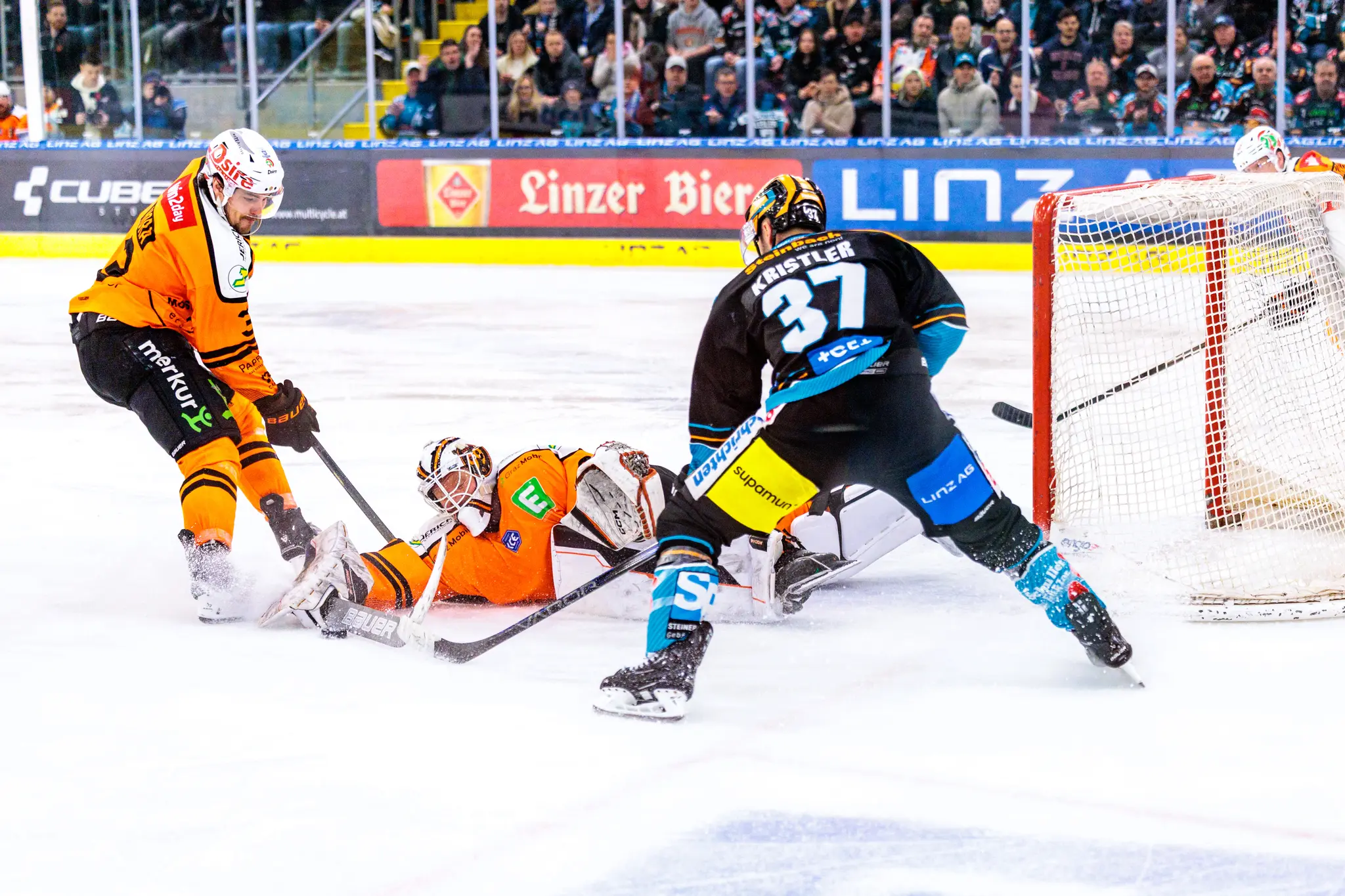 Eishockey-Spielszene mit einem Angreifer der Grazer 99ers beim Schuss auf das Tor, dem Torhüter in orange bei einer Parade und einem Black Wings Linz Spieler in dunkler Ausrüstung, der um den Puck kämpft 