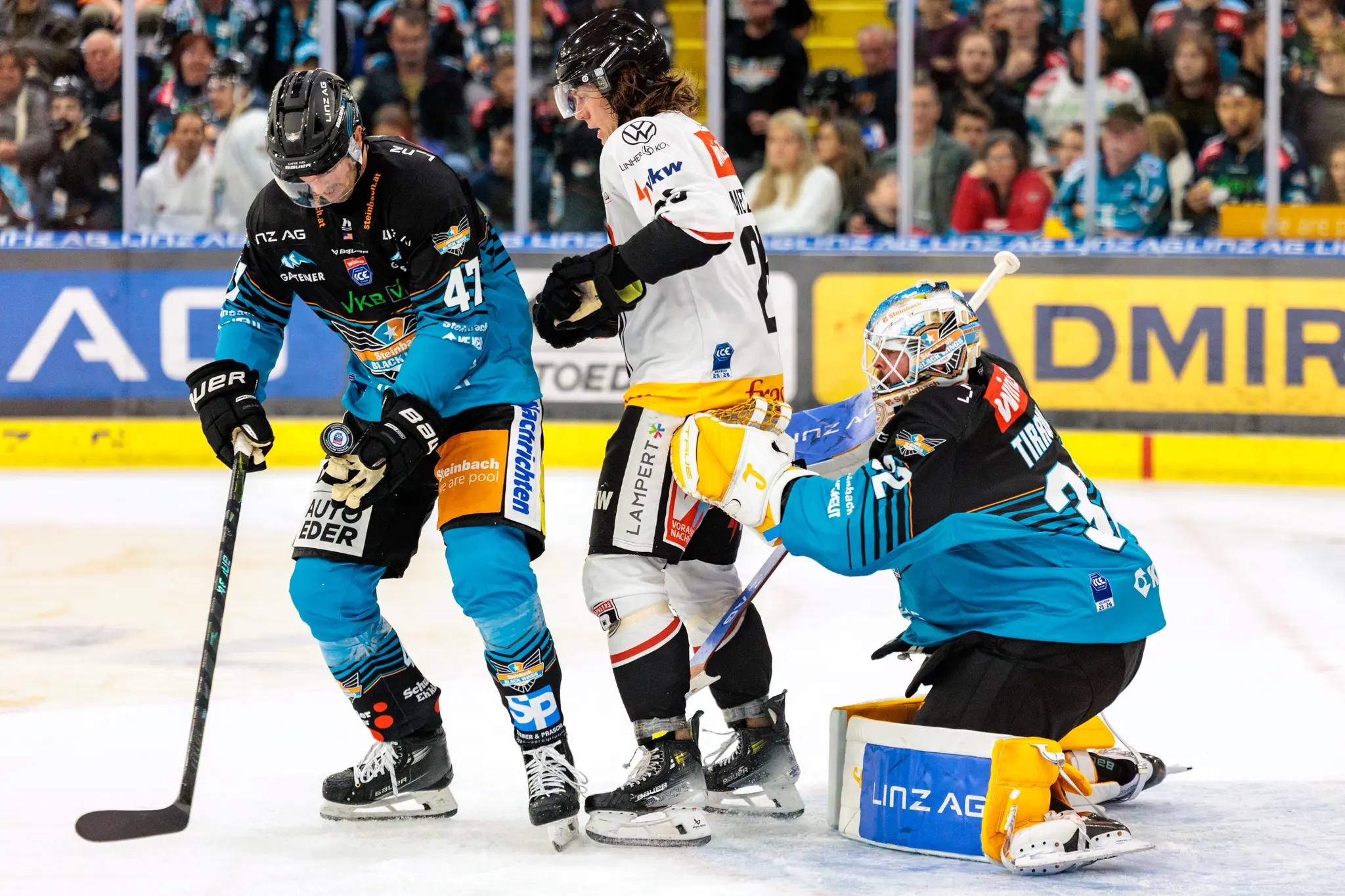 Eishockey-Torhüter Rasmus Tirronen von den Steinbach Black Wings Linz kniet zur Abwehr vor seinem Tor, während Julian Metzler und Logan Roe um den Puck kämpfen