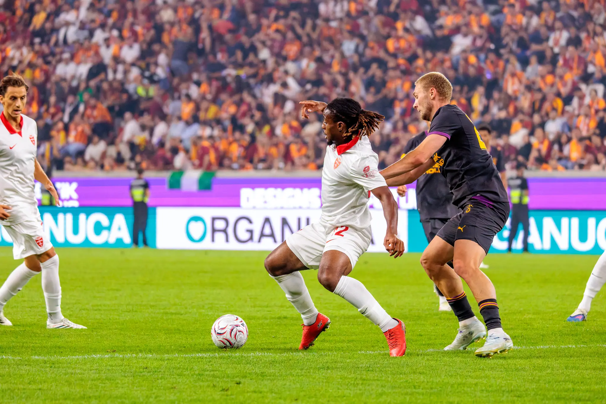 Monza-Spieler mit roten Stollenschuhen dribbelt den Ball, ein Galatasaray-Spieler kommt von rechts herannahend.