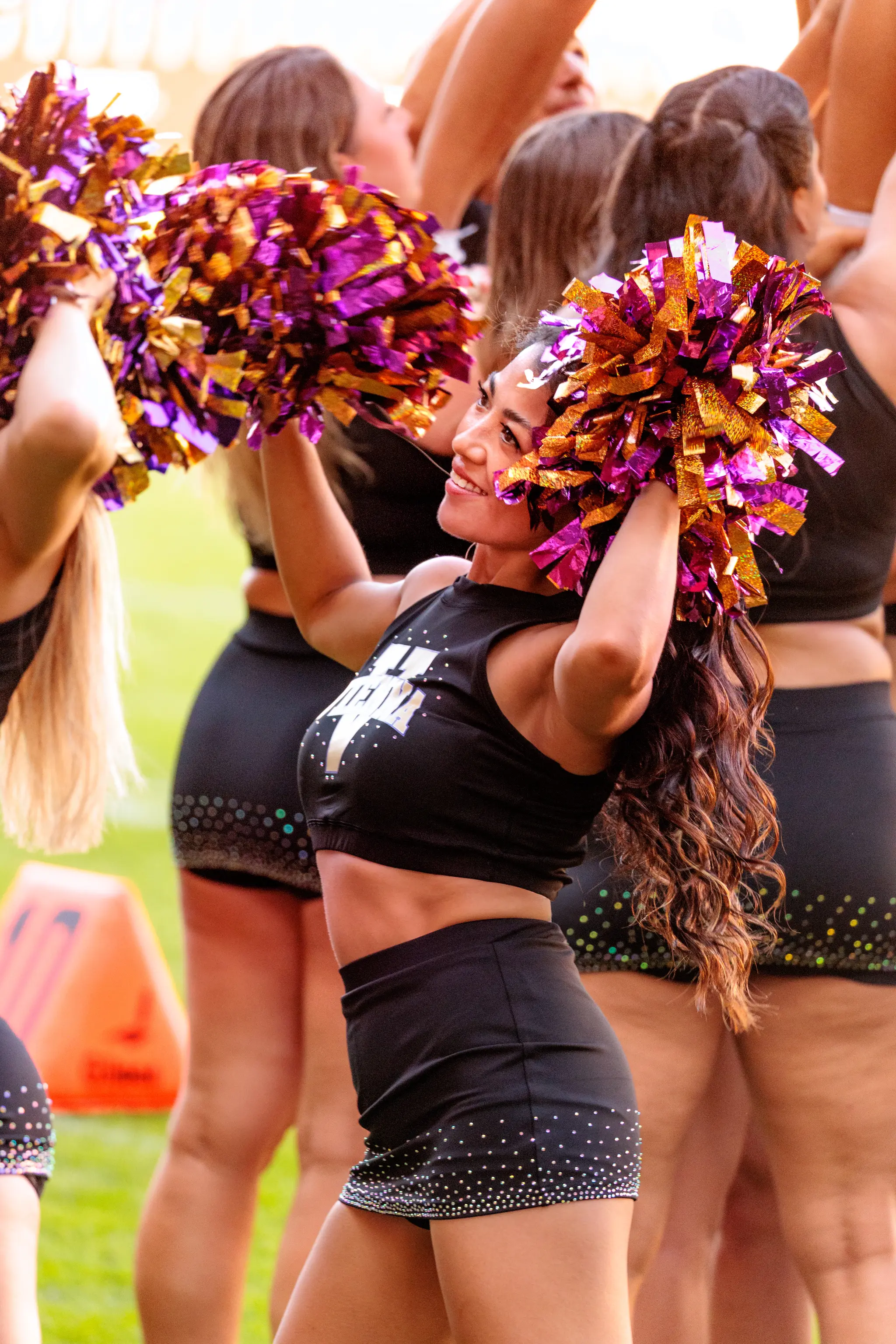 Cheerleaderin des Spirit Squad hebt beide violett-goldenen Pompons über den Kopf.