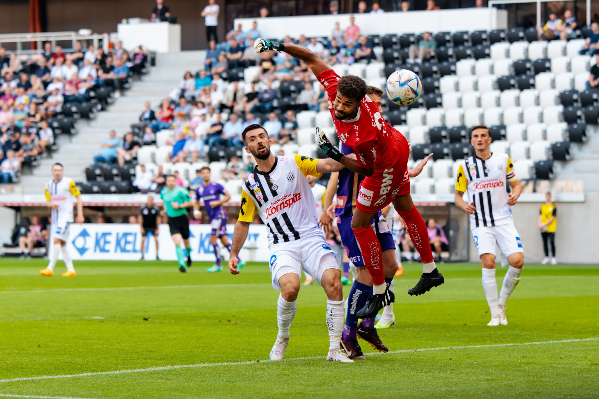 Ein LASK-Fußballspieler in schwarz-weißem Trikot und ein Gegner in rotem Dress duellieren sich in der Luft um den Ball während eines Spiels im Stadion