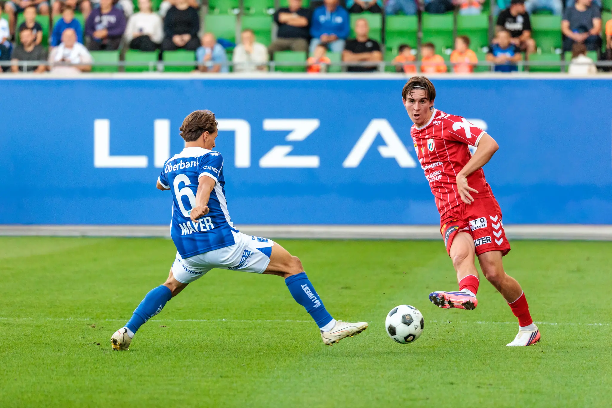 Jonas Mayer vom FC Blau-Weiß Linz geht im Mittelfeld in den Zweikampf gegen einen Welser Spieler; im Hintergrund ist die Linz-AG-Bande zu sehen.
