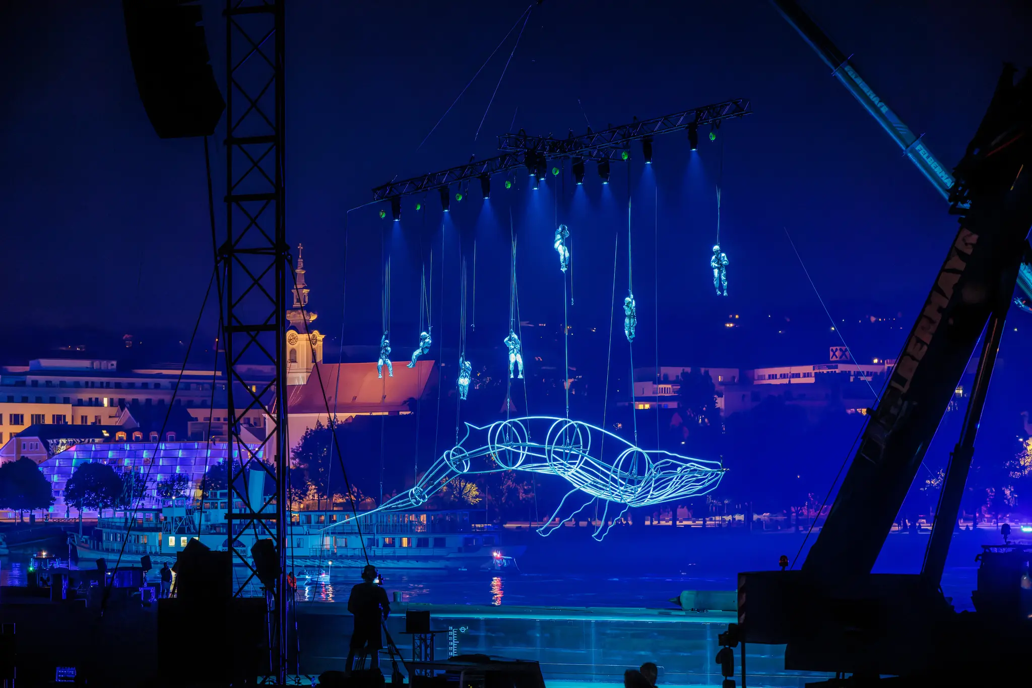 Abendliche Freiluft-Lichtshow mit schwebenden Akrobaten und einem leuchtenden Walskelett über einem Gewässer, vor der abendlichen Kulisse von Linz während der Klangwolke 2024 