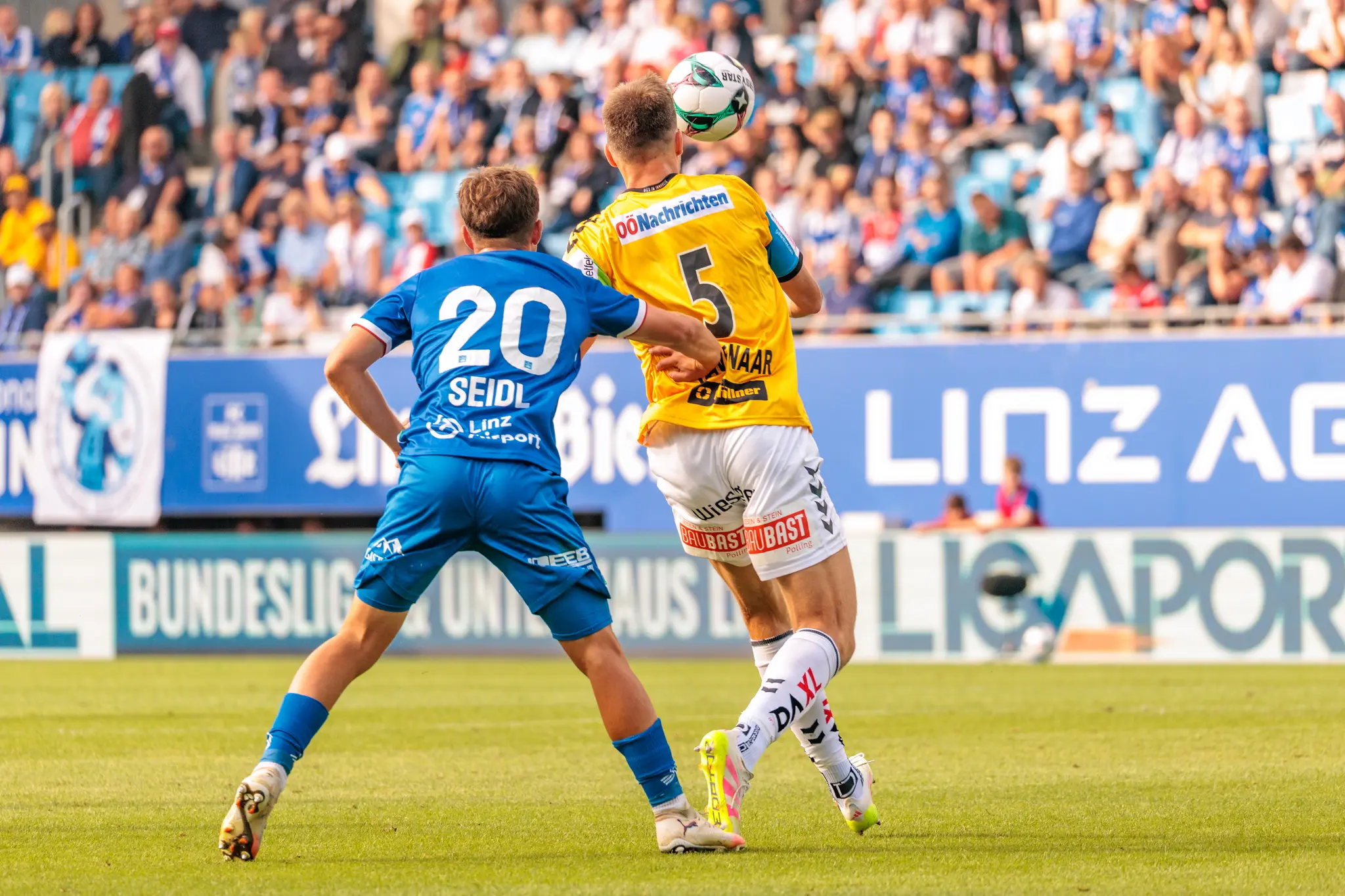 Ein Fußballspieler im gelben Trikot köpft den Ball in einem Duell mit einem Spieler im blauen Trikot, beobachtet von einer verschwommenen Zuschauermenge auf der Tribüne