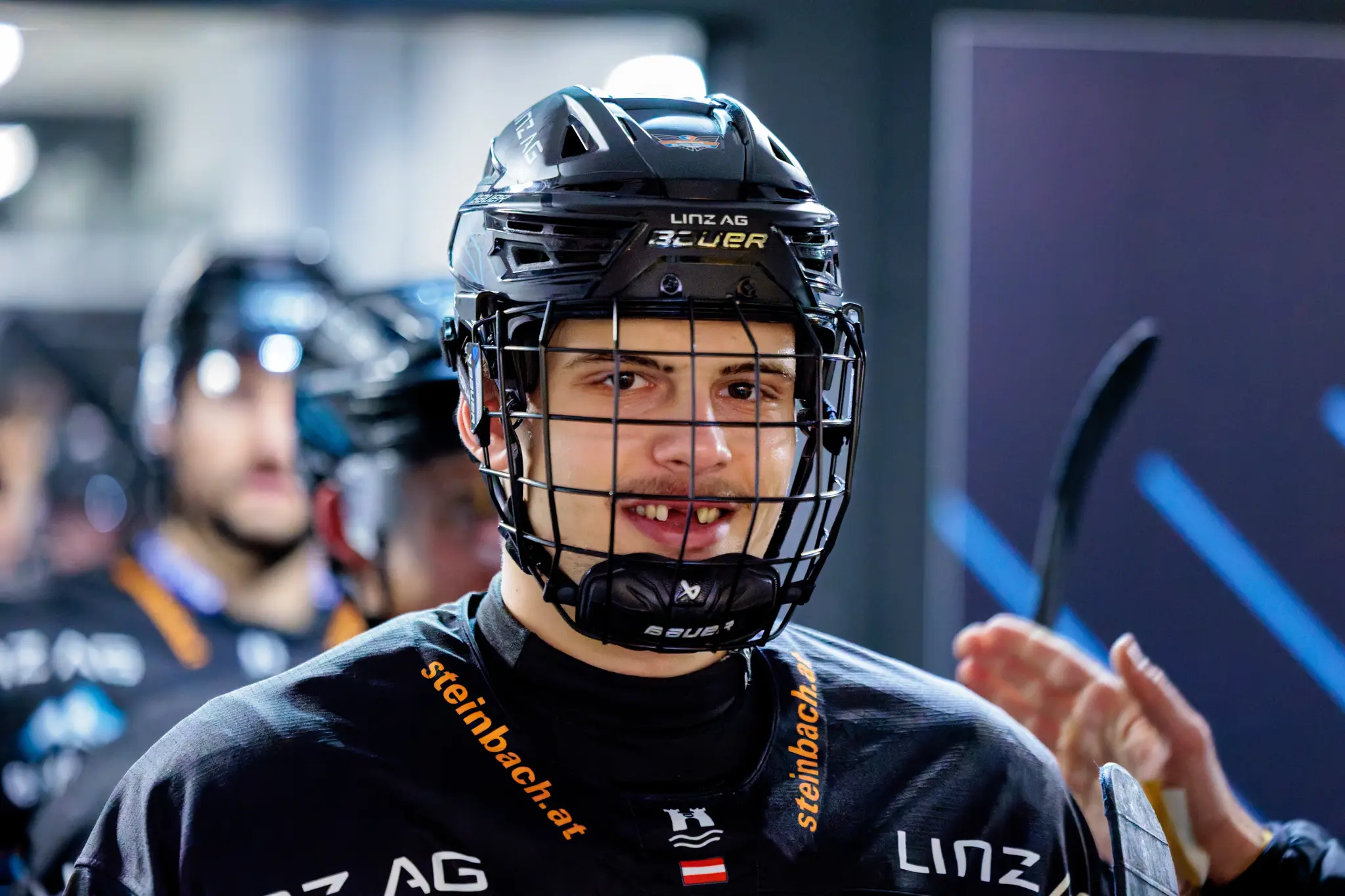 Porträt eines jungen Eishockeyspielers der Steinbach Black Wings Linz, der mit seinem schwarzen Helm und Gitter leicht in die Kamera lächelt