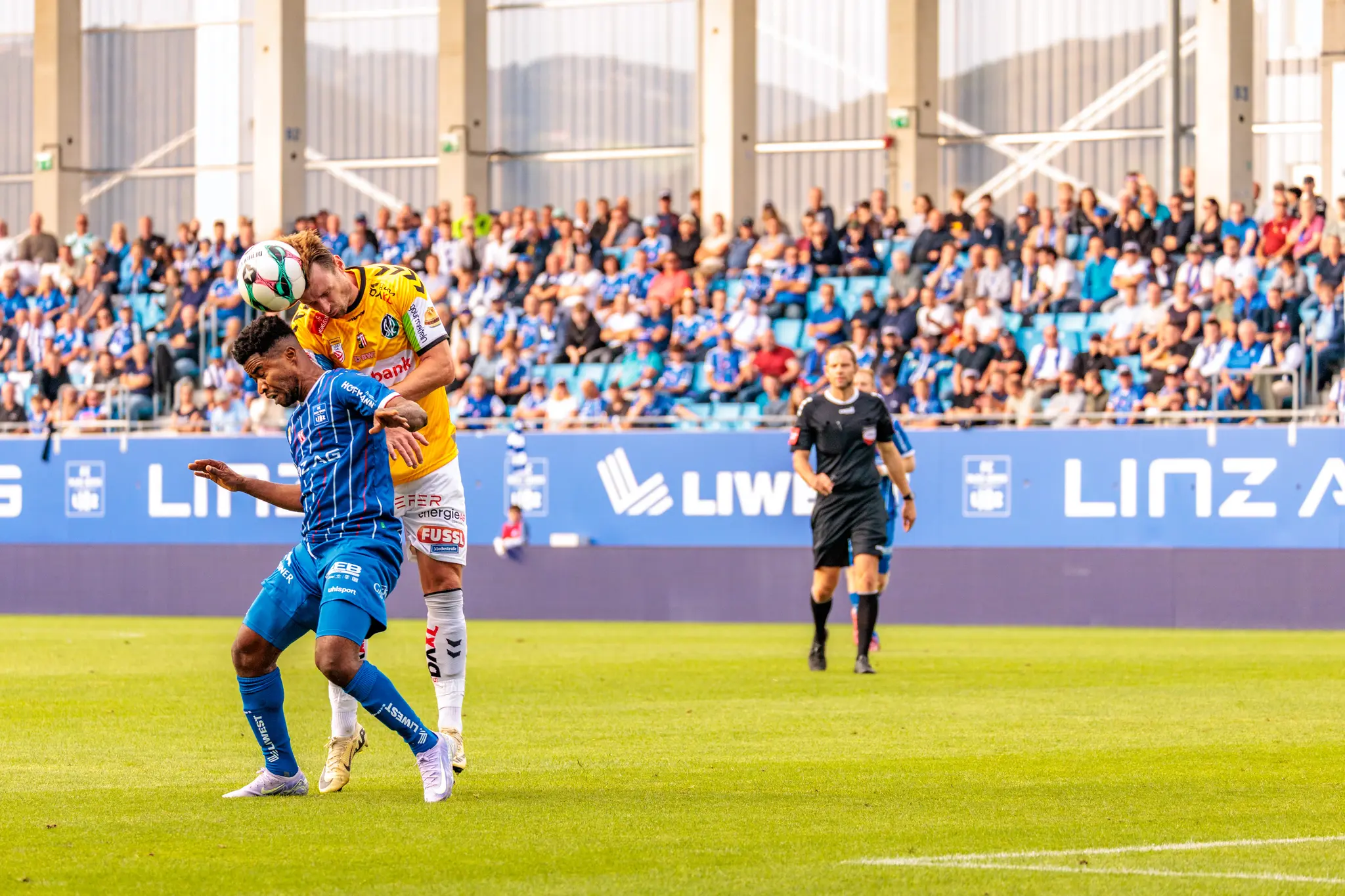 Zwei Fußballspieler liefern sich im Mittelfeld ein Kopfballduell, während ein Schiedsrichter und zahlreiche Zuschauer im Hintergrund das intensive Spiel verfolgen