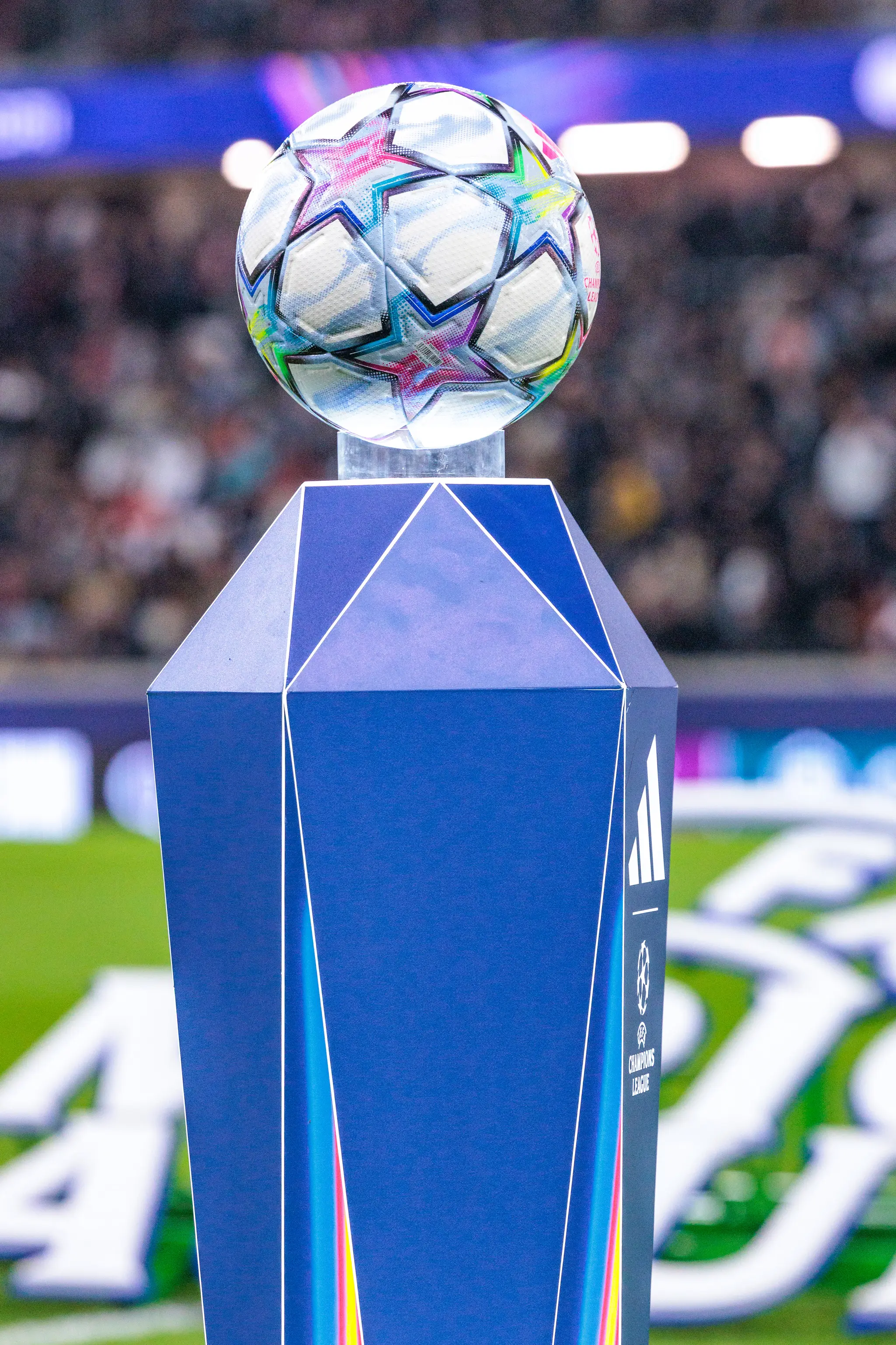 Der Champions-League-Spielball liegt auf einem blauen Podest mit Turnier- und Herstellerlogo, dahinter verschwimmen die Zuschauerränge.