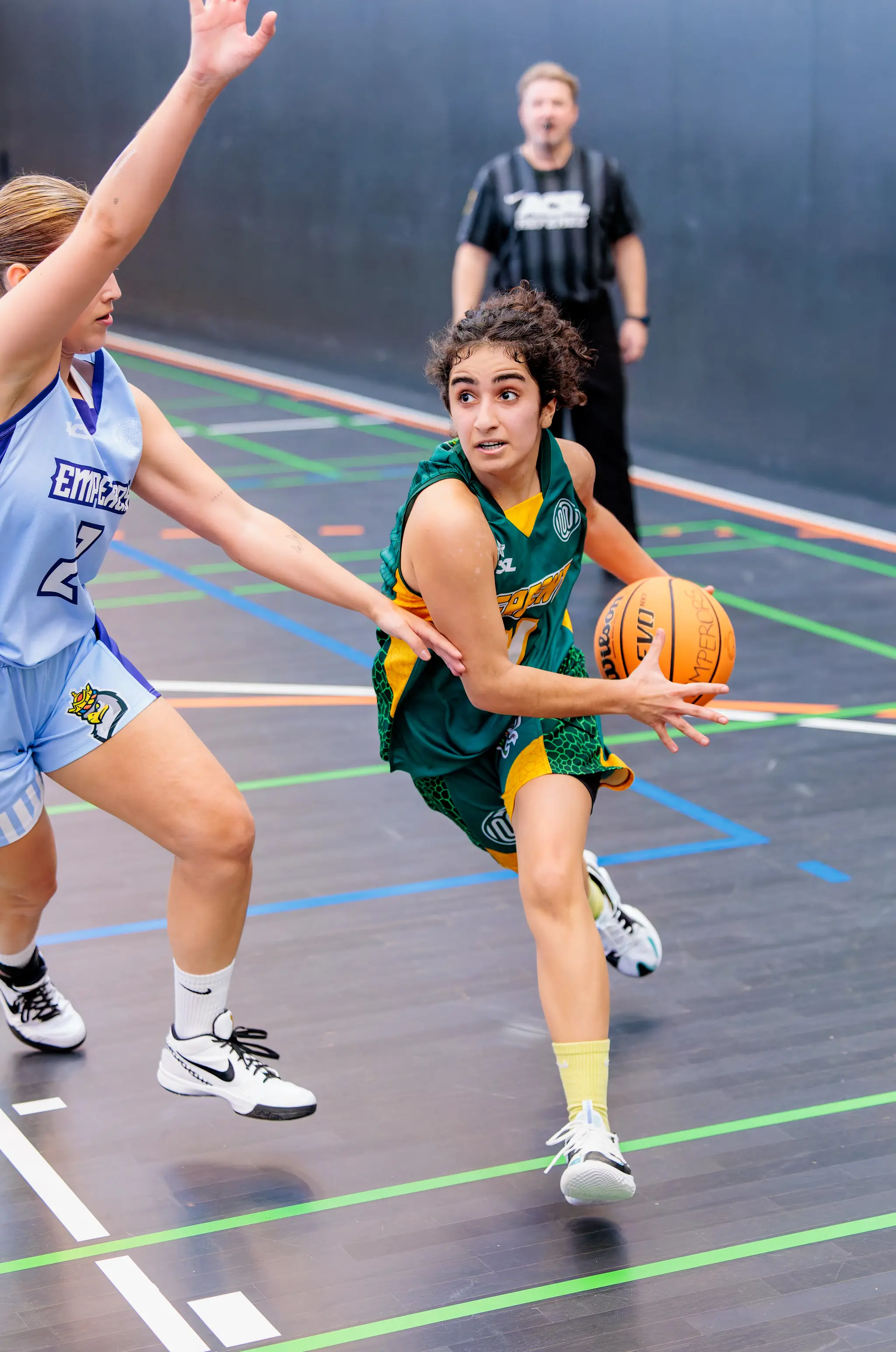 Eine Basketballspielerin im grünen Trikot dribbelt dynamisch den Ball, während eine Gegenspielerin im hellblauen Trikot verteidigt 