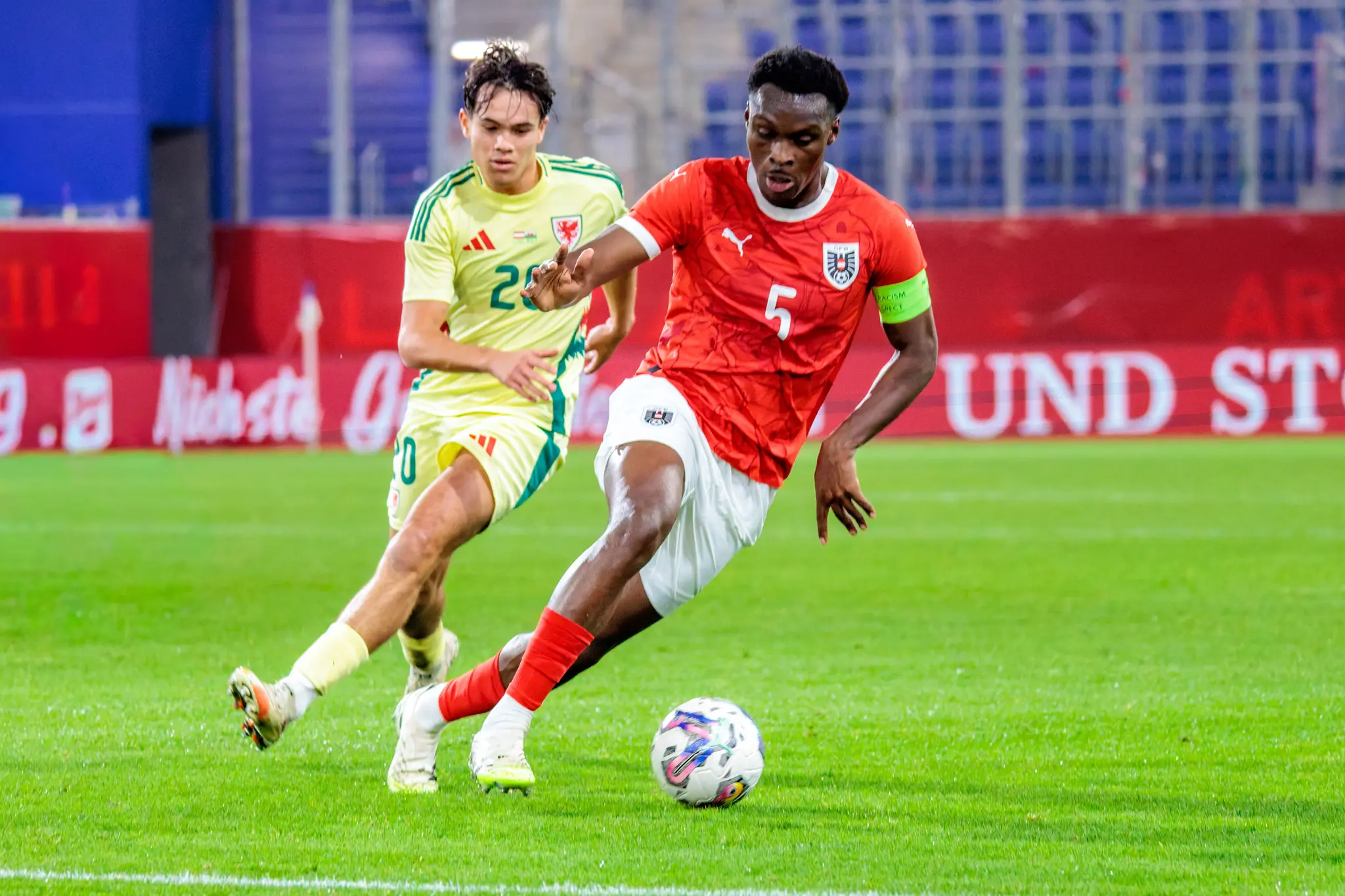 Samson Baidoo (Nummer 5, Österreich) im roten Trikot führt den Ball im Zweikampf mit Gabriele Biancheri (Nummer 20, Wales) im gelben Trikot