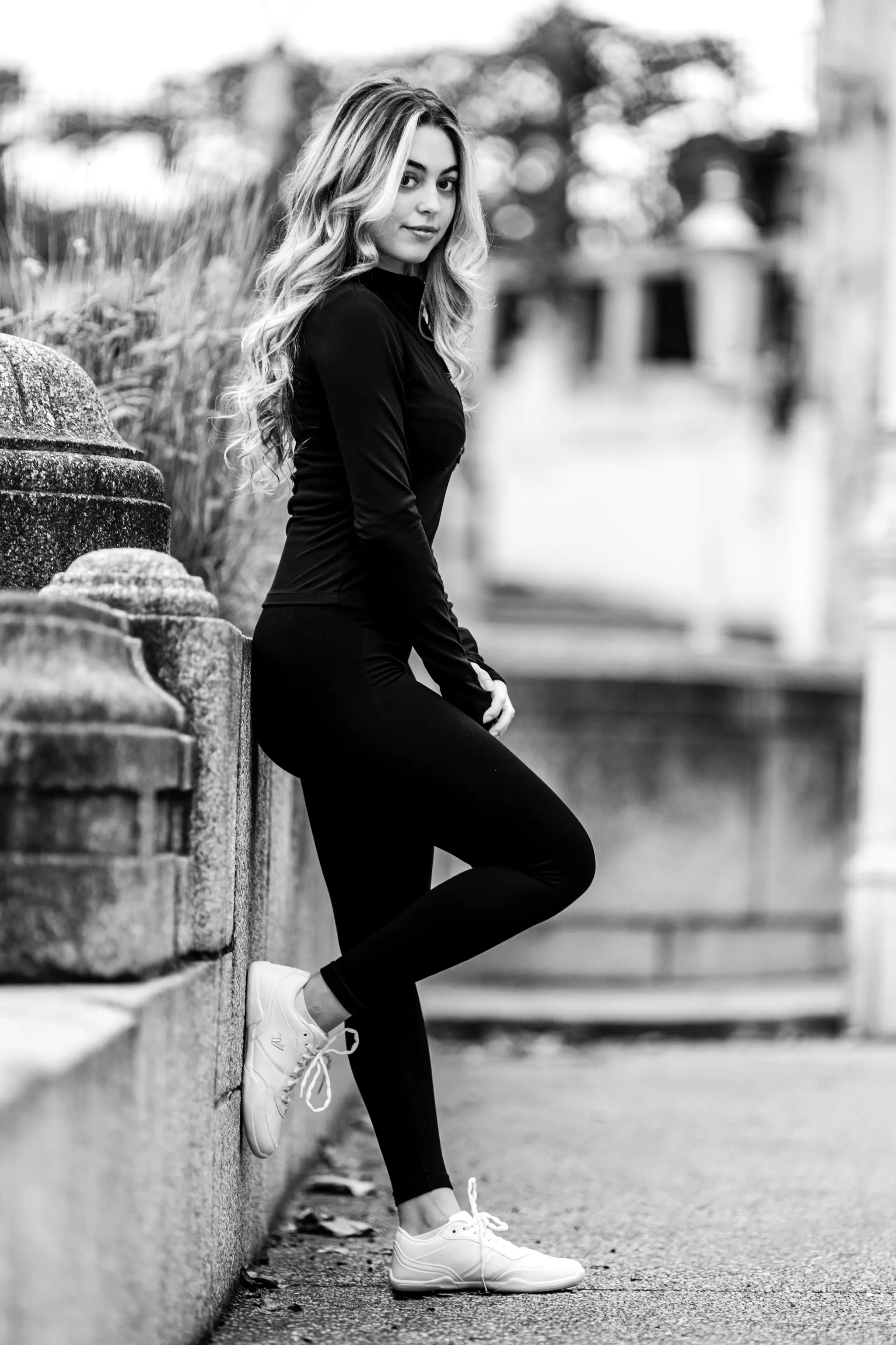 Schwarz-Weiß-Ganzkörperporträt von Model Valentina, die in einem sportlichen schwarzen Outfit an einer Steinmauer im Stadtpark lehnt und in die Kamera blickt