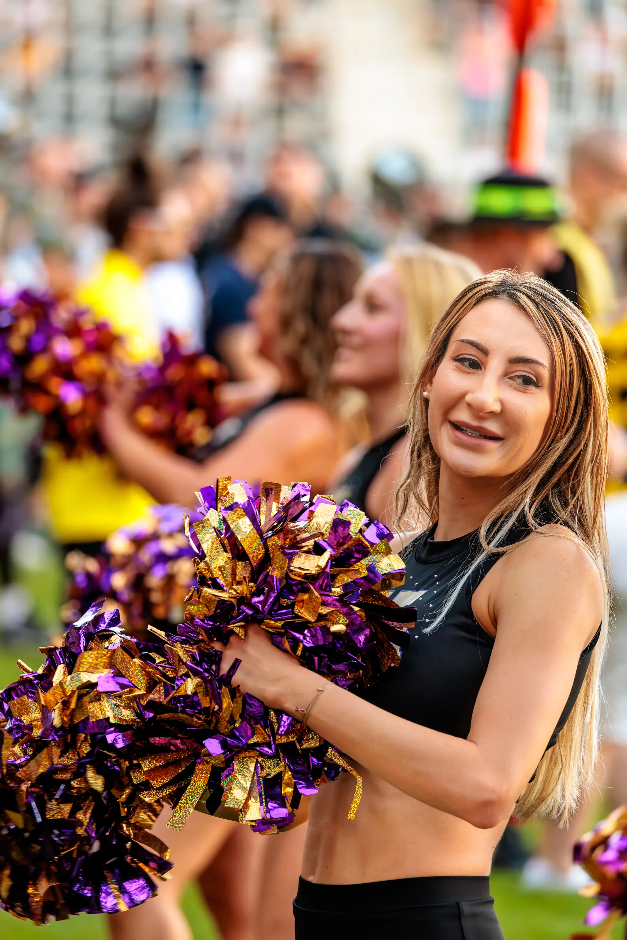 Lächelnde Cheerleaderin des Spirit Squad bei einer Tanzbewegung mit violett-goldenen Pompons.