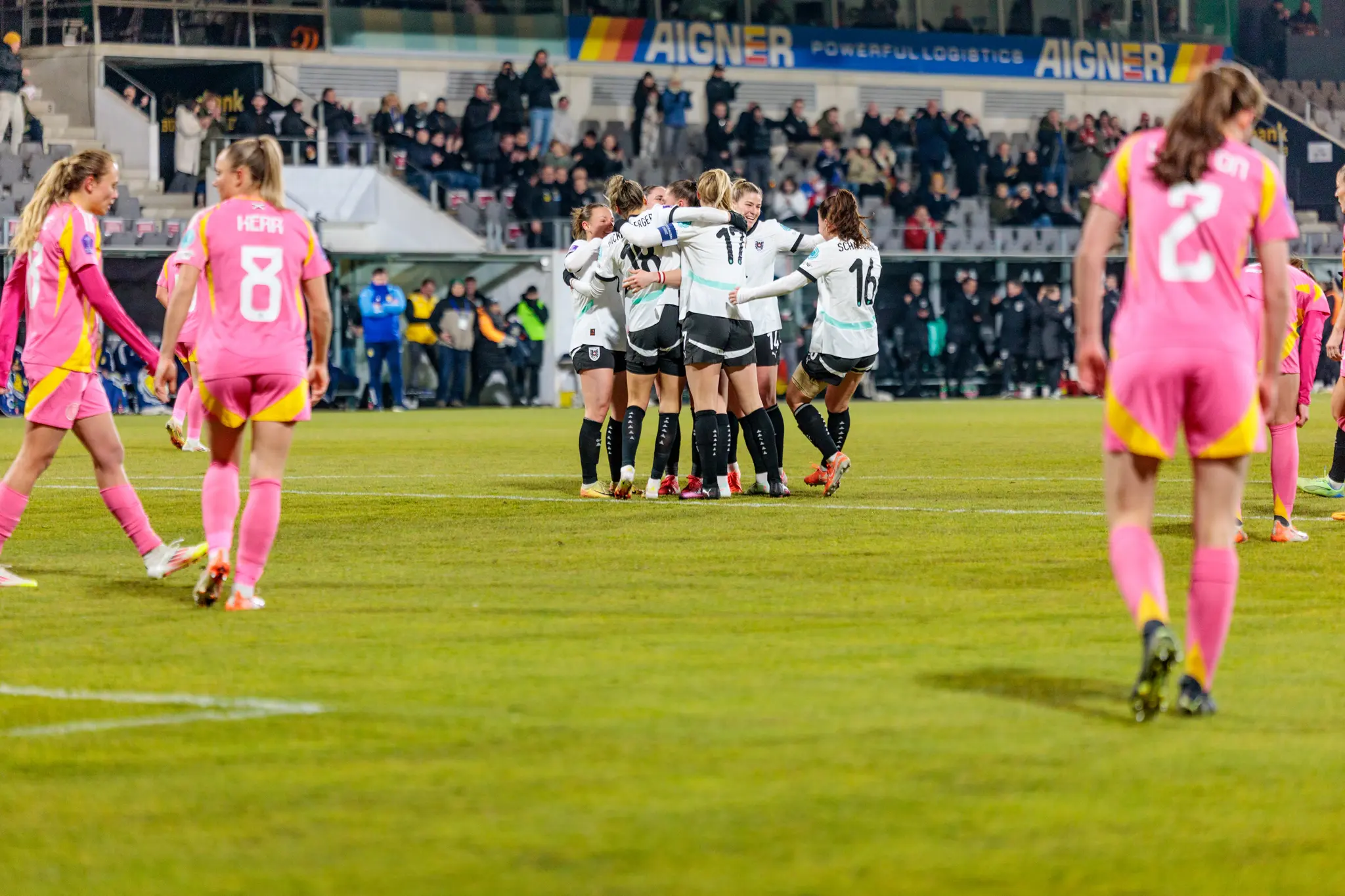 Jubelnde österreichische Fußballerinnen in weißen Trikots umarmen sich auf dem Feld, umgeben von Gegnerinnen in pinken Trikots und Zuschauern im Stadion
