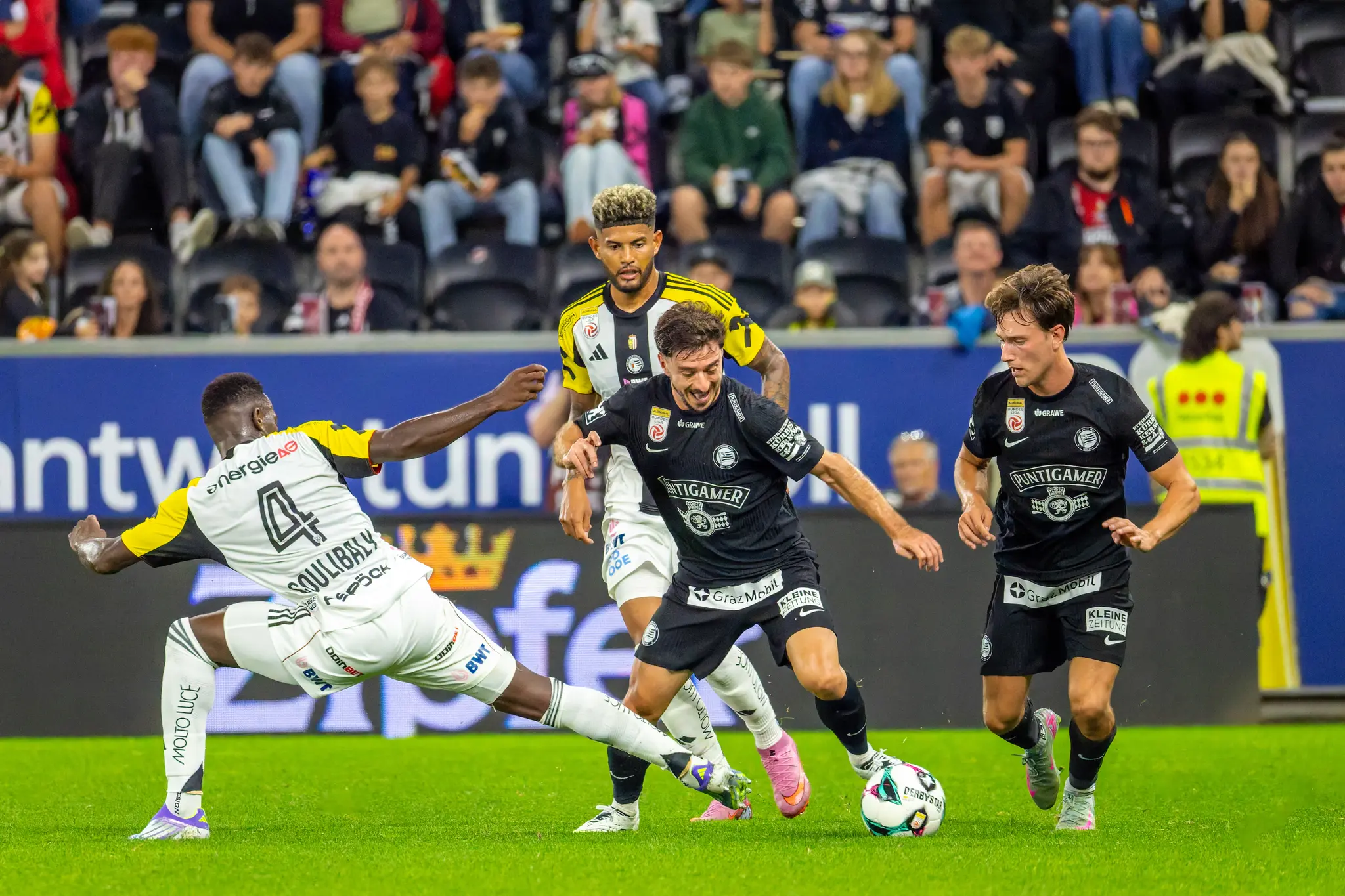 Vier Fußballspieler sind in einen intensiven Zweikampf um den Ball verwickelt, wobei ein Spieler von Sturm Graz den Ball am Fuß führt und ein Spieler des LASK eine Grätsche ausführt