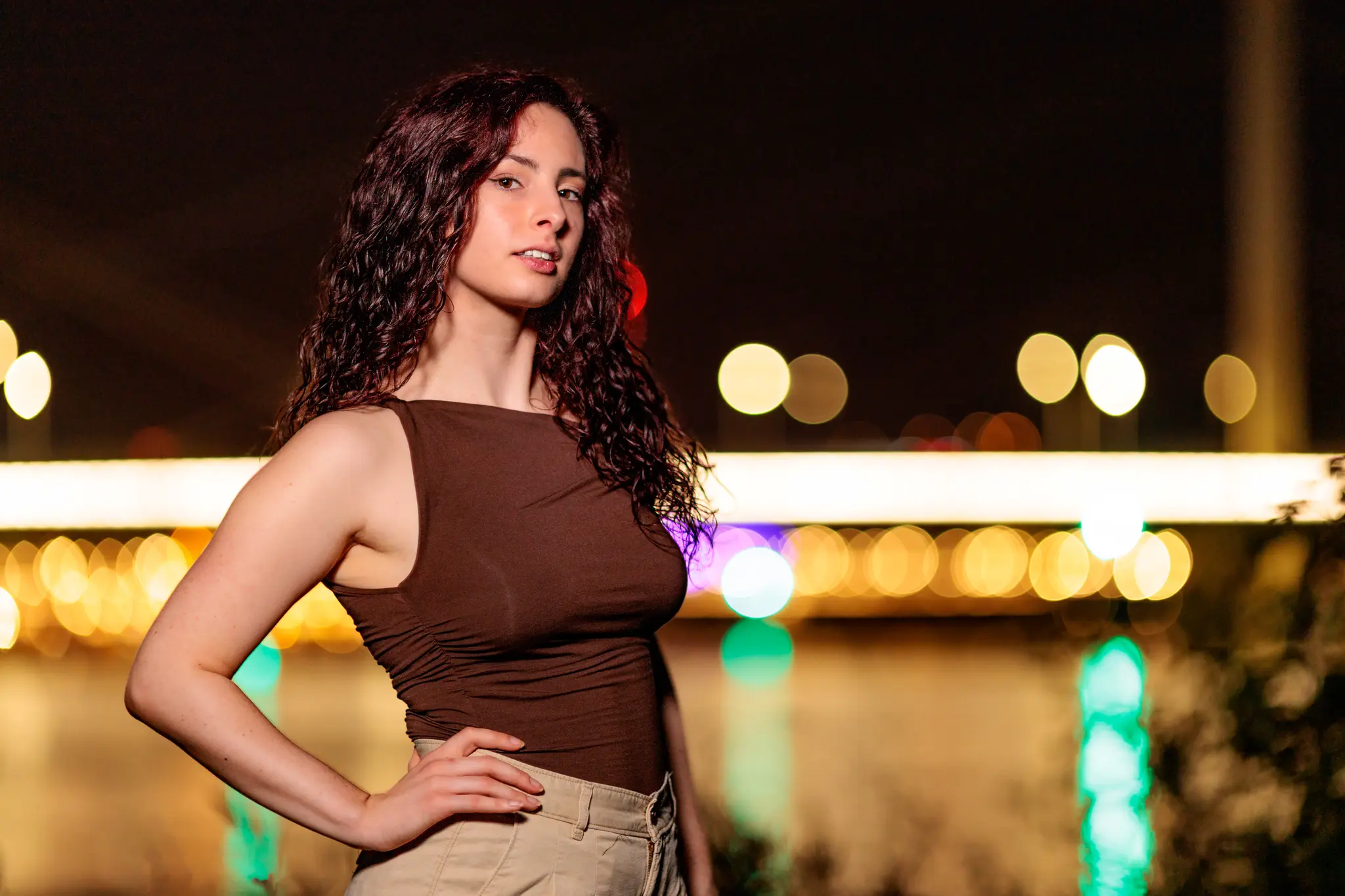 Ein Portrait von Nicole mit lockigem Haar in einem braunen Oberteil, selbstbewusst stehend vor dem nächtlichen Linz mit seinen funkelnden Bokeh-Lichtern