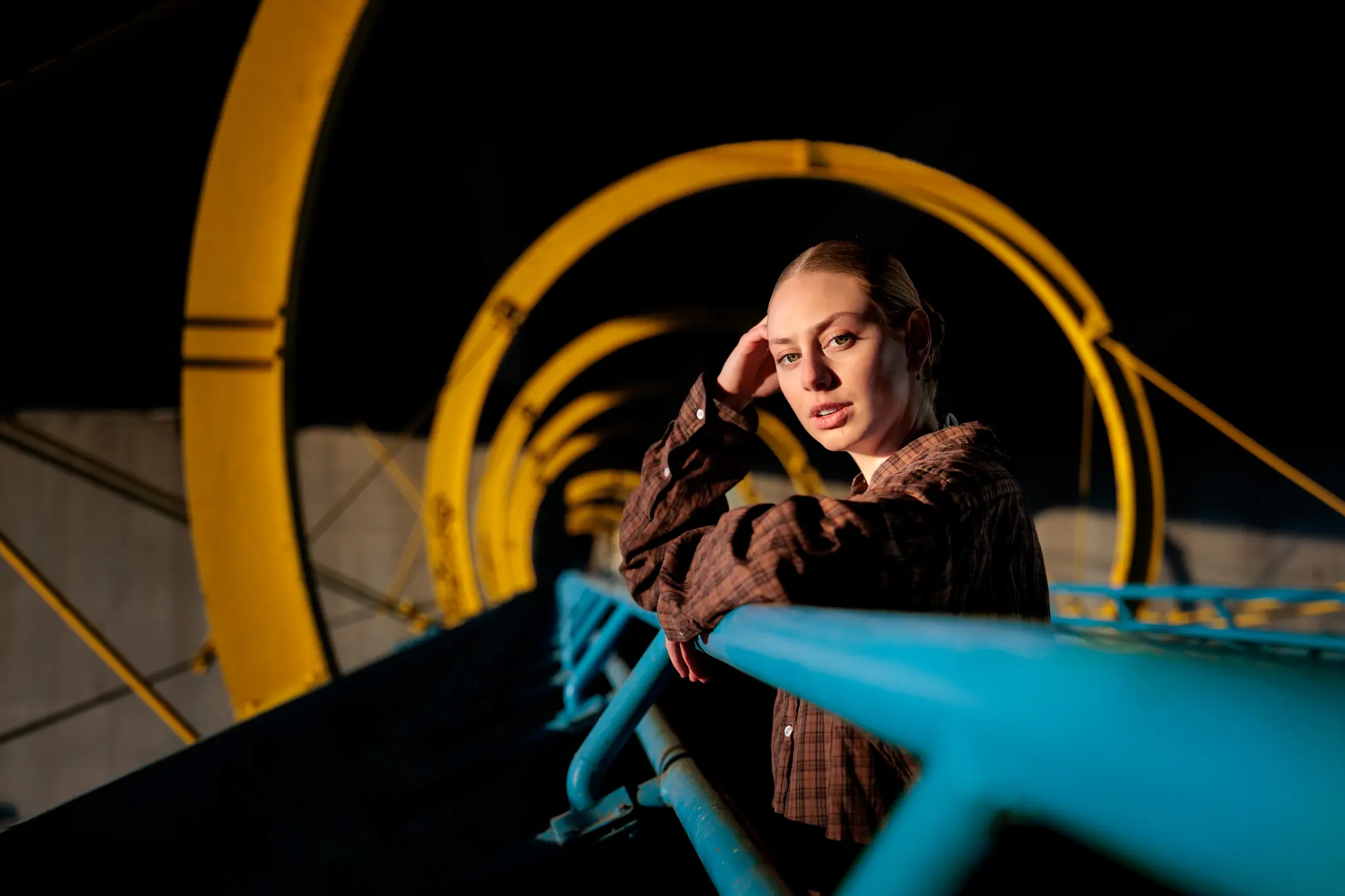 Eine junge Frau mit blonden Haaren lehnt an einem blauen Geländer in einem geometrischen Tunnel aus gelben Bögen. 