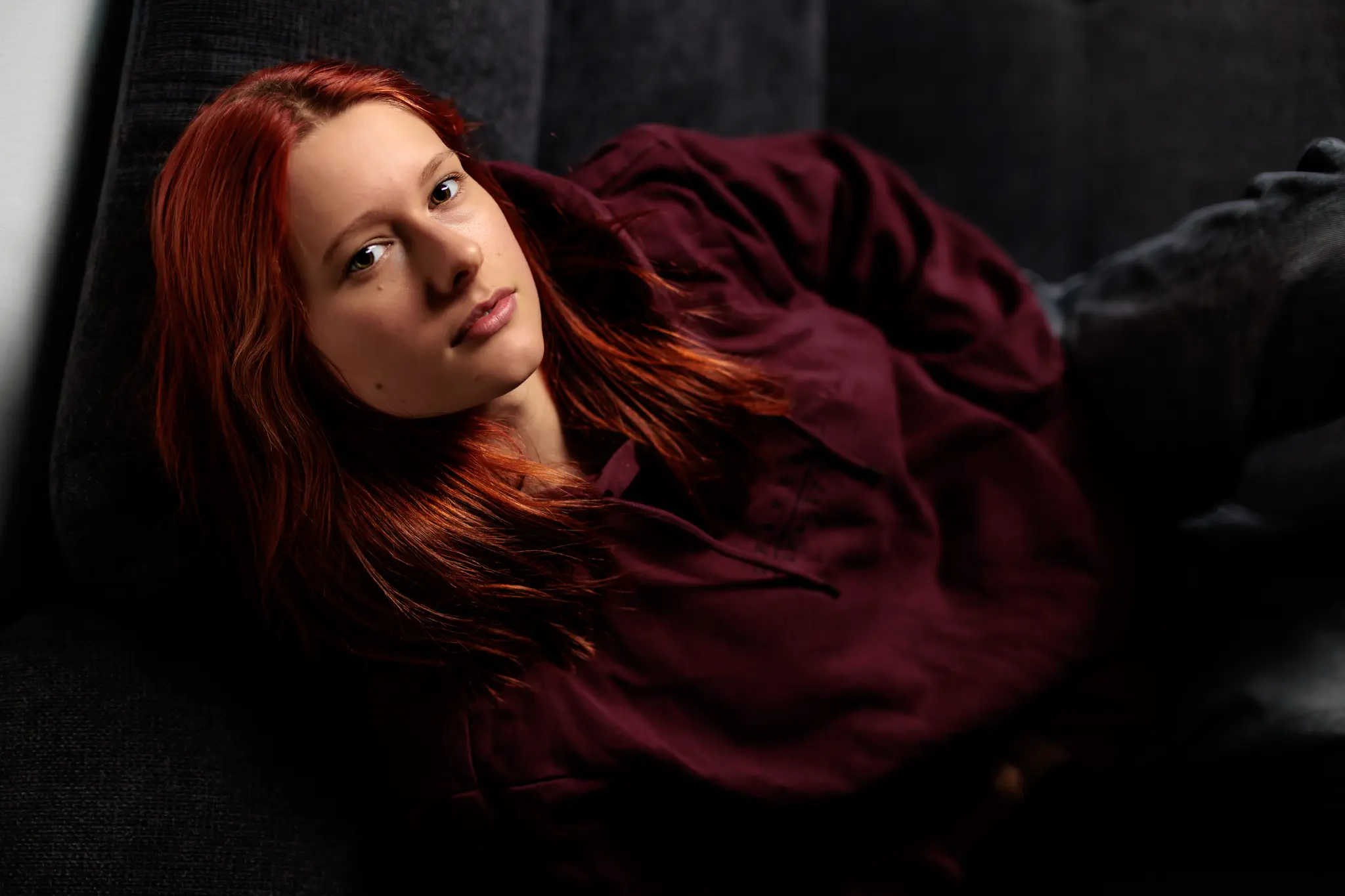 Sarah mit markanten roten Haaren blickt in einem bordeauxfarbenen Hoodie bei einem atmosphärischen Beauty-Home-Shooting intensiv in die Kamera