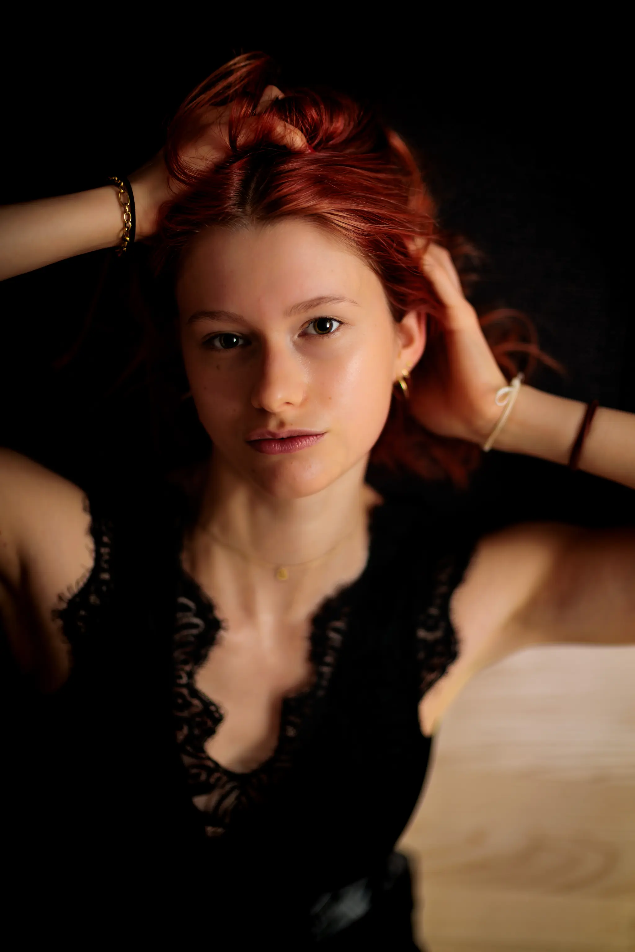 Ausdrucksstarkes Portrait von Sarah mit markanten roten Haaren und den Händen im Haar, die in einem eleganten schwarzen Spitzentop intensiv in die Kamera blickt