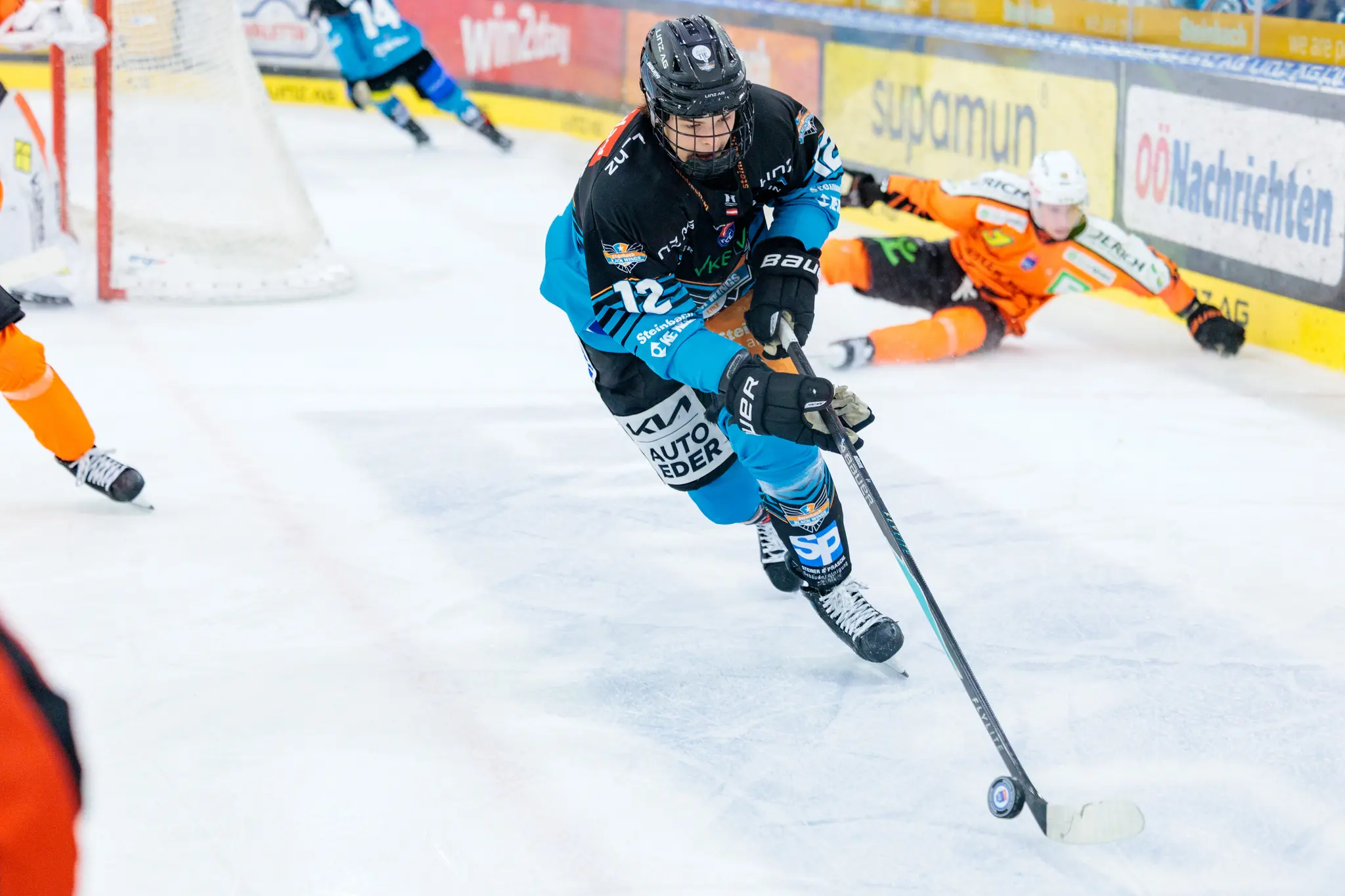 Ein Eishockeyspieler der Black Wings Linz in schwarz-blauen Trikot greift mit dem Schläger an, während ein Gegenspieler der Grazer 99ers in orange im Hintergrund auf das Eis stürzt
