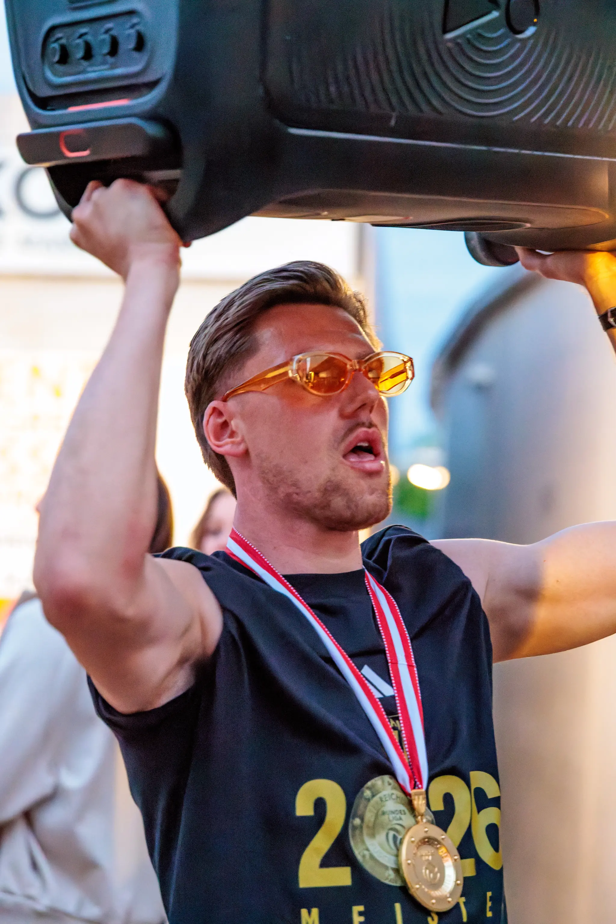 Ein LASK-Spieler mit Sonnenbrille und SiegerMedaille hebt eine Musikbox während der Meisterfeier in Linz hoch
