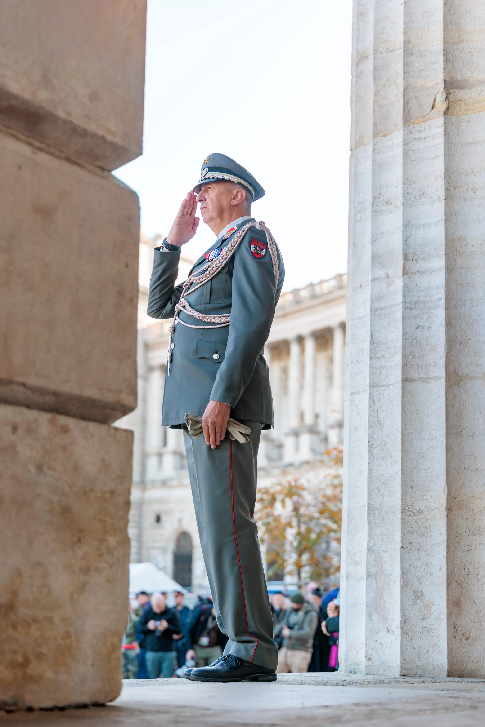 Ein Offizier in Ausgehuniform salutiert zwischen mächtigen Steinsäulen, im Hintergrund stehen Besucherinnen und Besucher am Heldenplatz.