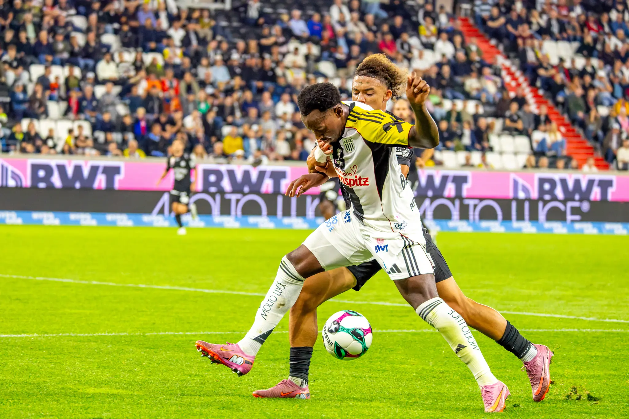 Zwei Fußballspieler in unterschiedlich farbigen Trikots ringen eng um den Ball auf dem Spielfeld vor einer gefüllten Zuschauertribüne
