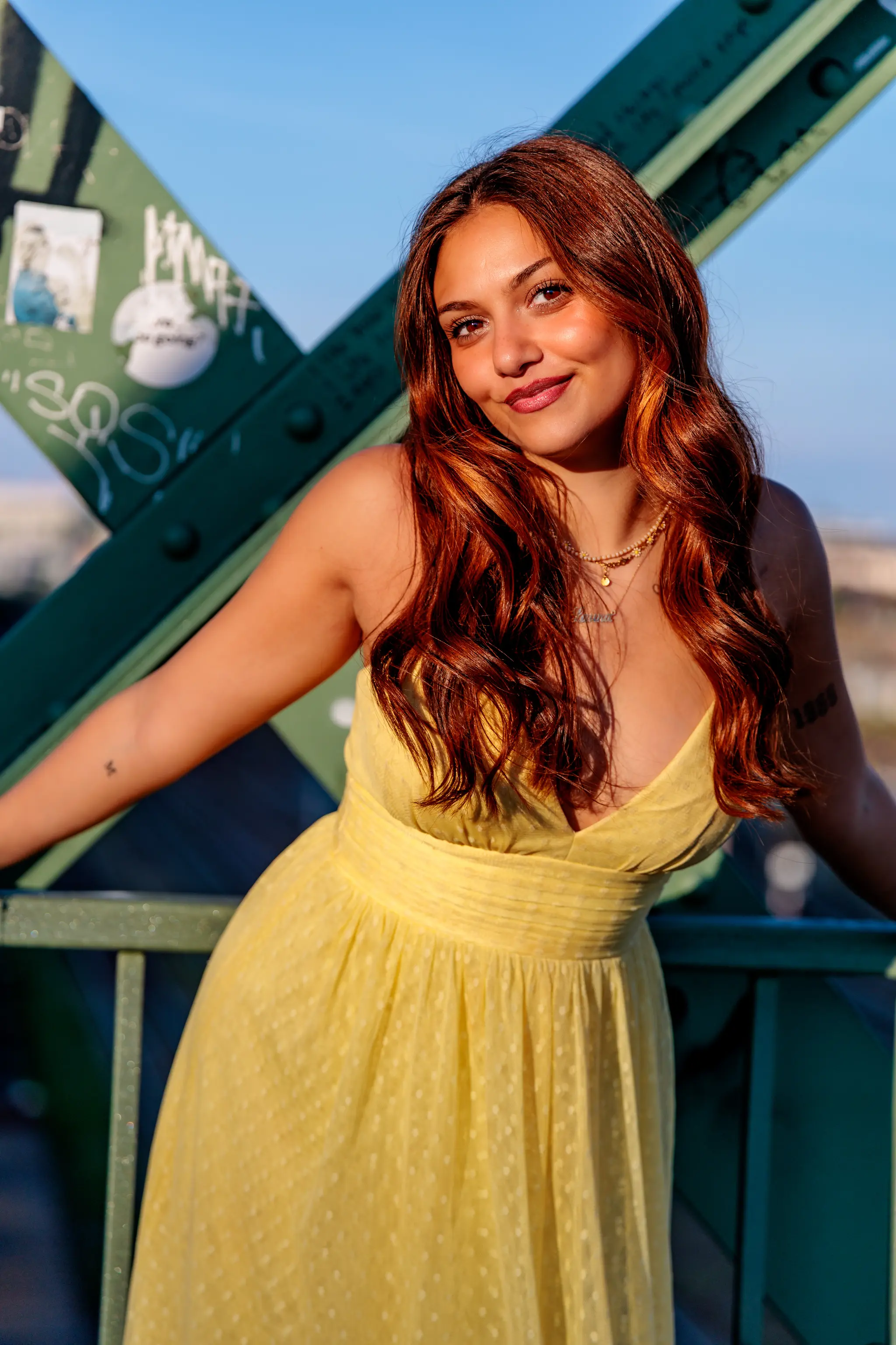 Lachende junge Frau mit roten Haaren in gelbem Sommerkleid vor grüner Brücke im Sonnenschein. 