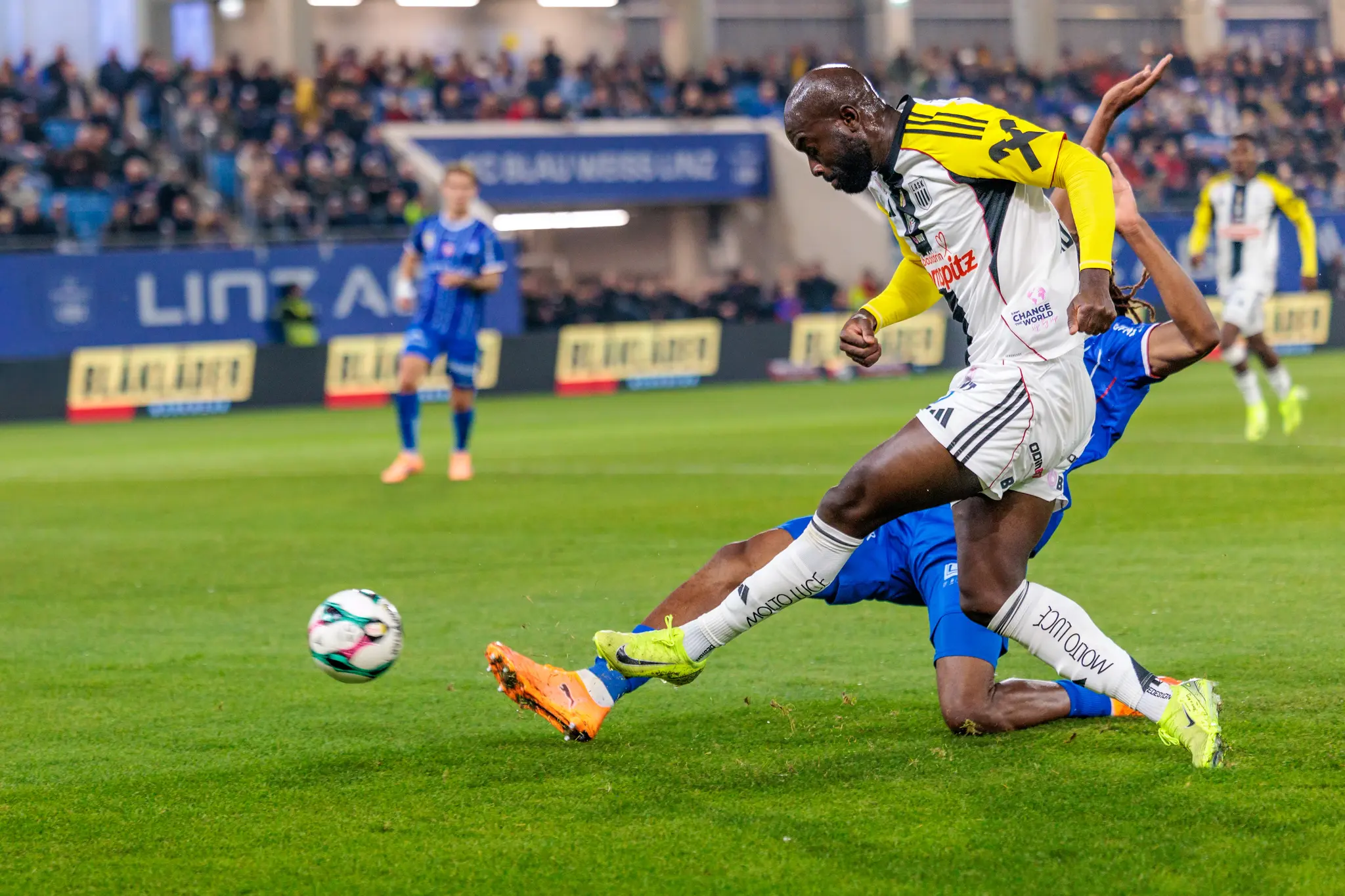 Moses Adeniran im gelb-weißen LASK-Trikot gibt einen Torschuss ab, während ein Spieler von Blau-Weiß Linz in blauem Trikot am Boden liegend blockt