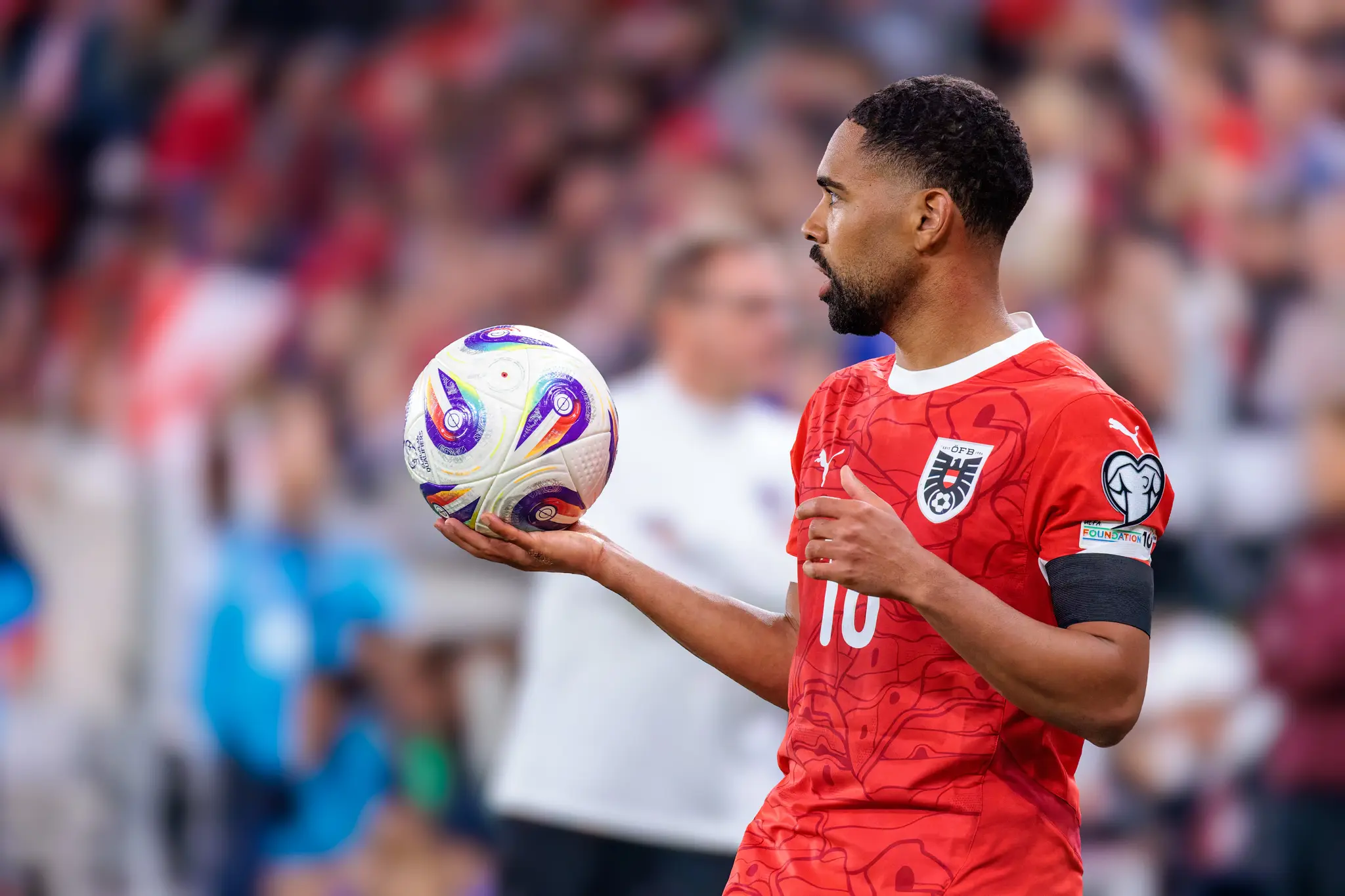 Der österreichische Nationalspieler Phillipp Mwene im roten Trikot mit der Rückennummer 10 hält einen offiziellen UEFA European Qualifiers Fußball in der Hand und blickt zu den Mitspielern