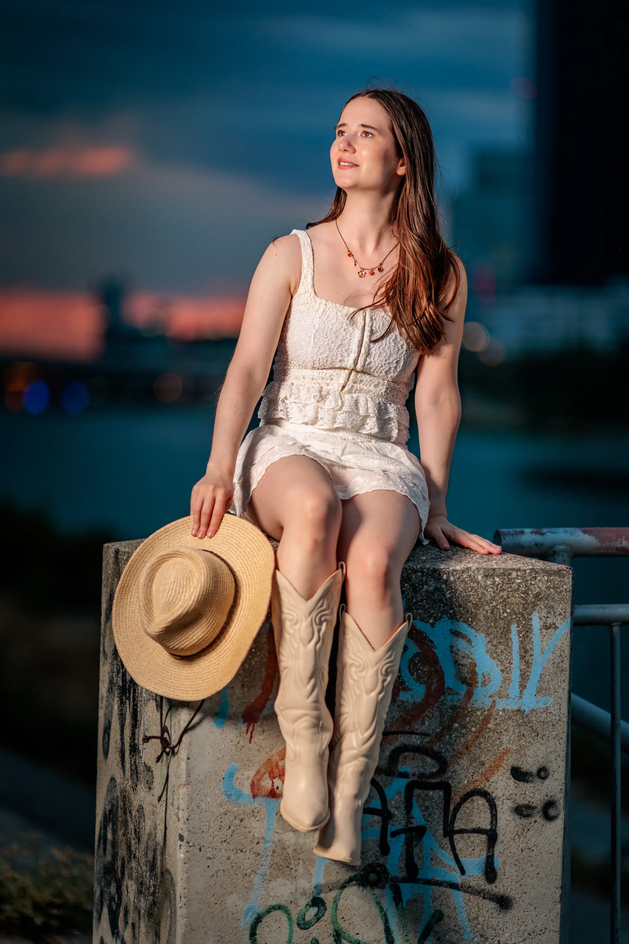 Hanna sitzt in der Abenddämmerung auf einem betonenen Graffiti-Pfeiler, die Cowboy-Stiefel an den Füßen und den Blick nach oben gerichtet.