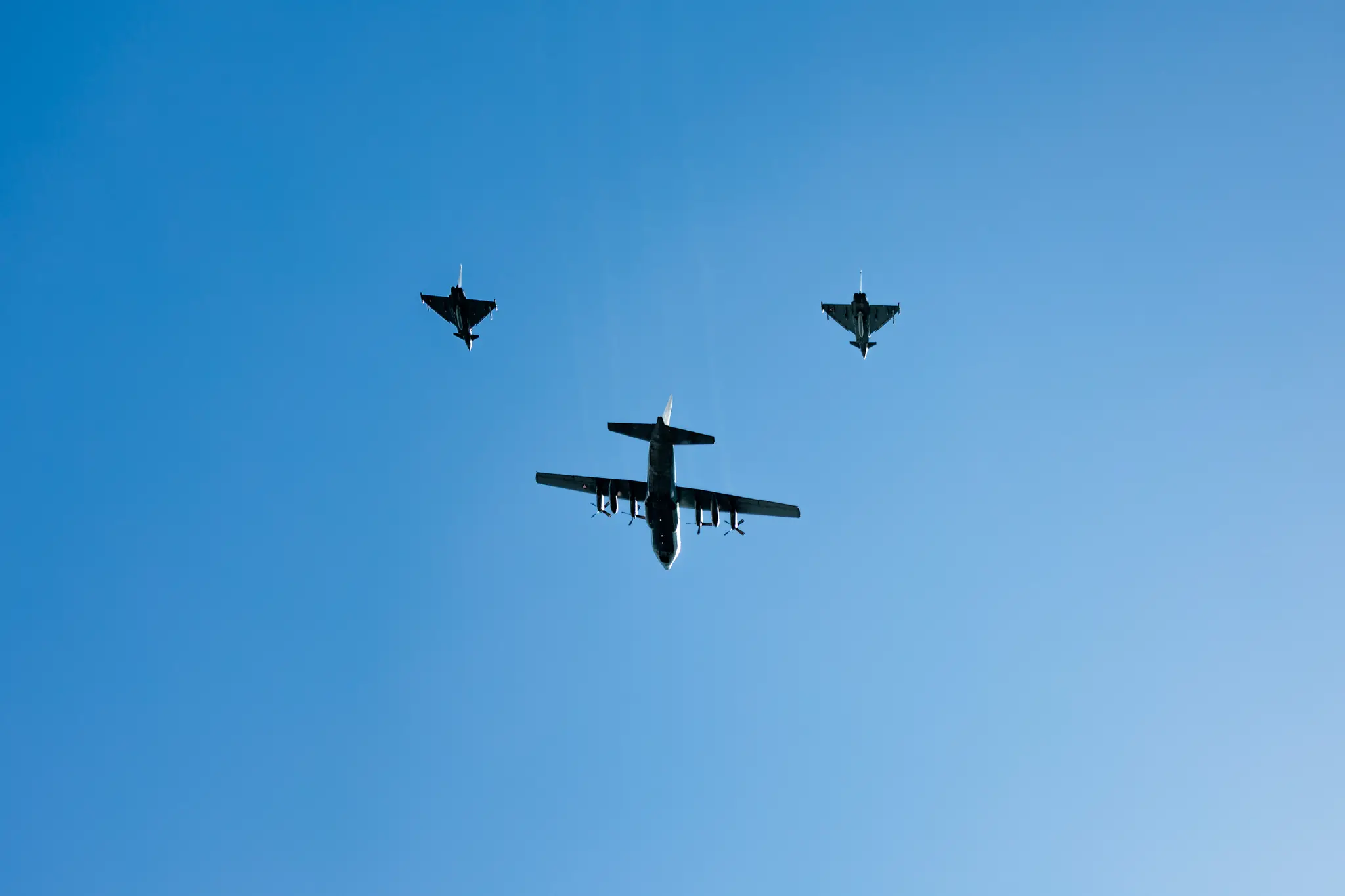 Eine Hercules-Transportmaschine fliegt in Formation mit zwei Eurofightern über den Heldenplatz, der Himmel leuchtet tiefblau.