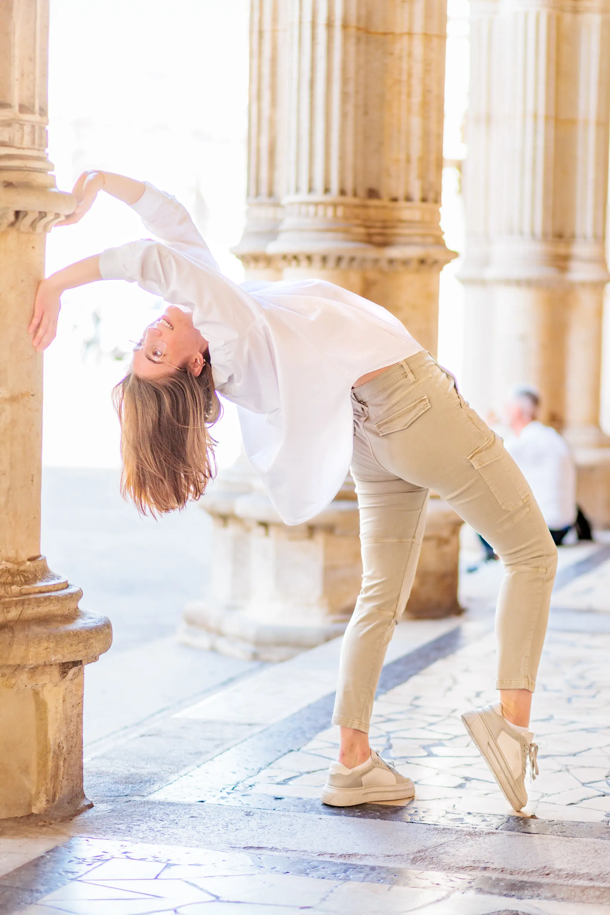 Elegante junge Frau in Weiß und Beige macht eine dynamische Rückbeuge an einer historischen Steinsäule der Wiener Oper 