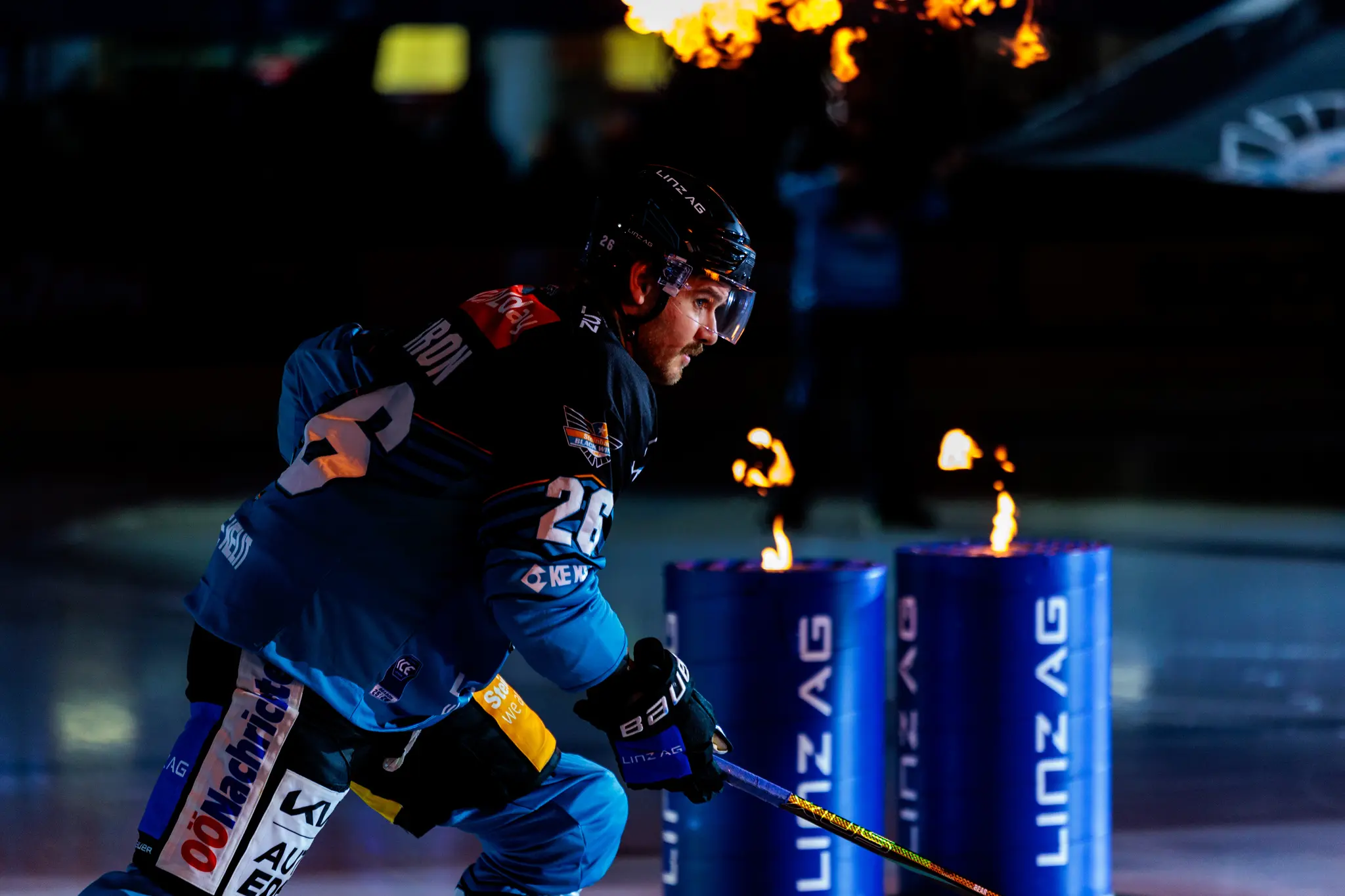 Eishockeyspieler Travis Barron in blauer und schwarzer Uniform gleitet beim Einlauf auf dem Eis, während Flammen aus Feuersäulen schlagen