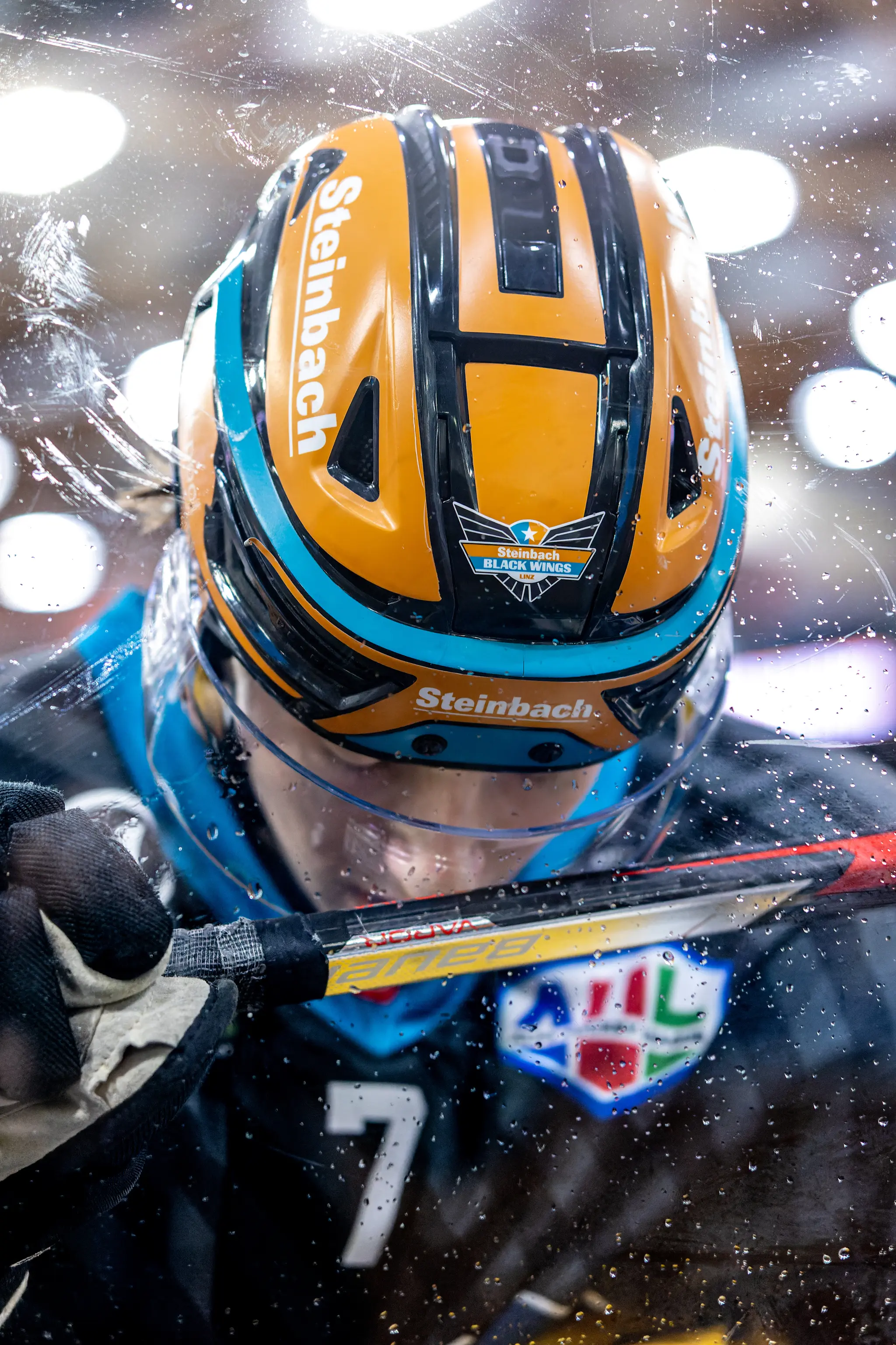 Ein Eishockey-Spieler der SteelWings mit orange-schwarzem Helm und der Nummer 7 auf dunklem Trikot ist durch eine nasse Plexiglasscheibe während eines Spiels zu sehen
