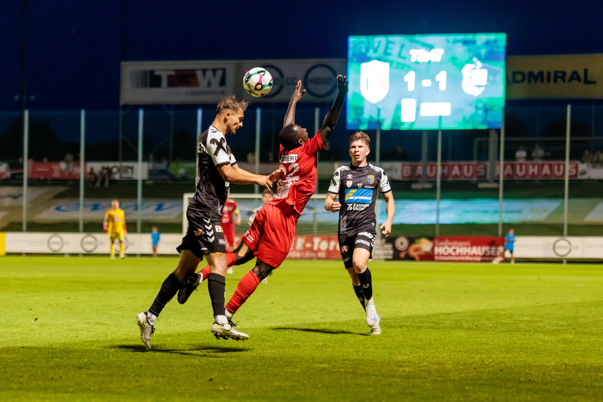 Spieler der SPG HOGO Wels und des SKN St. Pölten liefern sich einen dynamischen Luftzweikampf um den Ball auf dem Fußballfeld unter Flutlicht, während die Anzeigetafel ein 1:1 anzeigt
