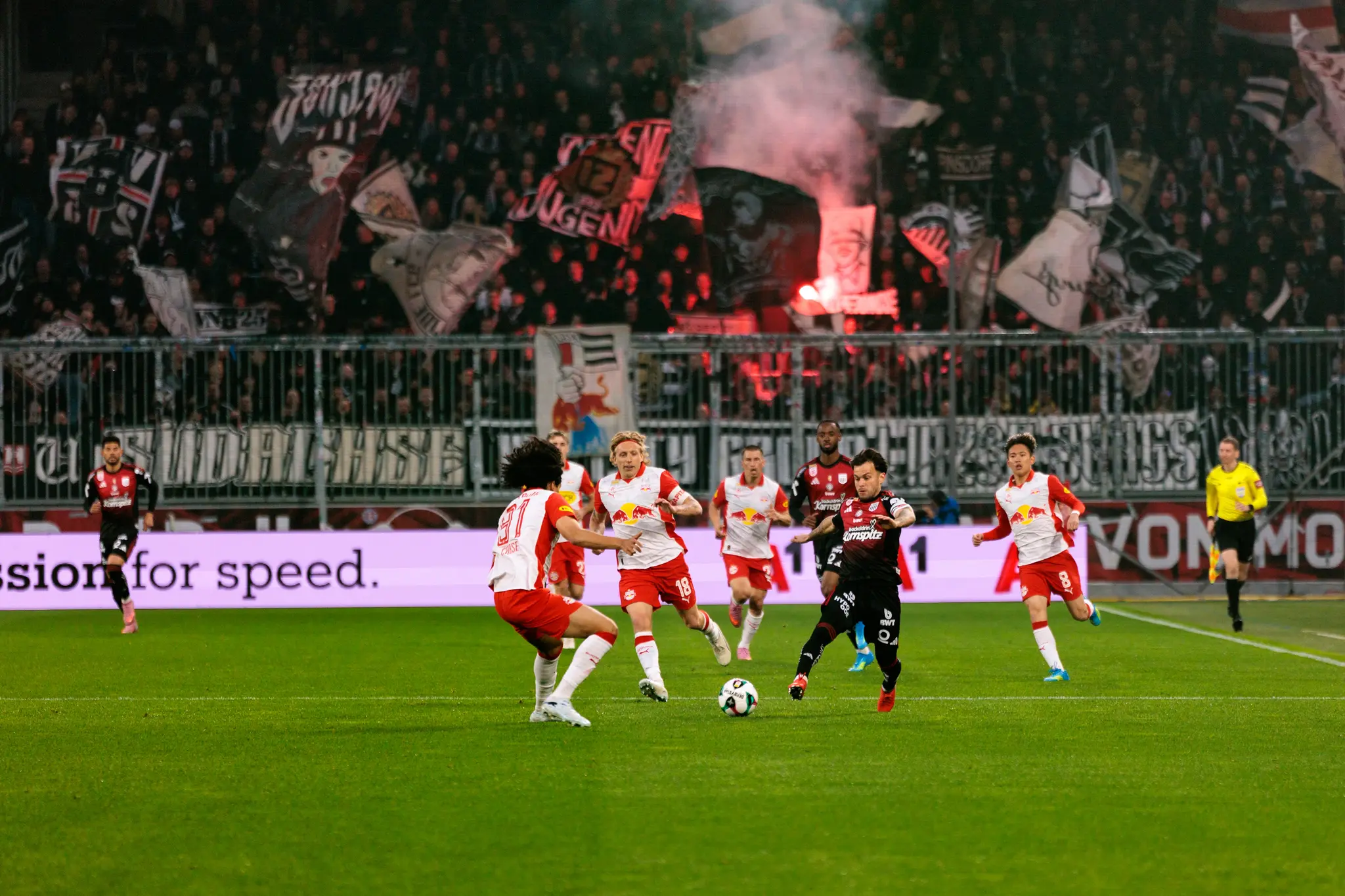 Ein Spieler des LASK führt den Ball im Dribbling gegen mehrere Verteidiger von Red Bull Salzburg vor einer mit Pyrotechnik und Fanbannern gefüllten Fankurve