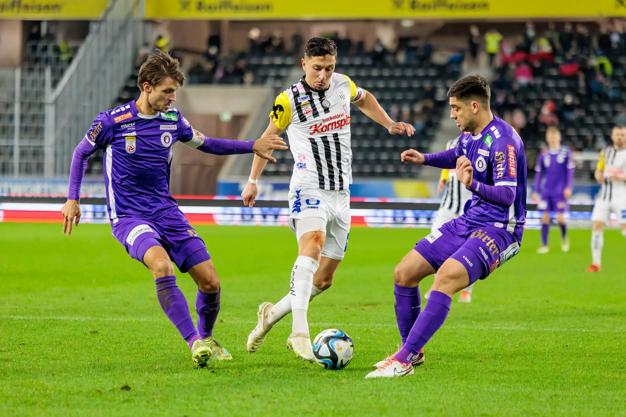 Ein Spieler im schwarz-weiß gestreiften Trikot im Zweikampf mit zwei Gegnern in lilafarbenen Trikots um den Fußball während einer Bundesliga-Partie